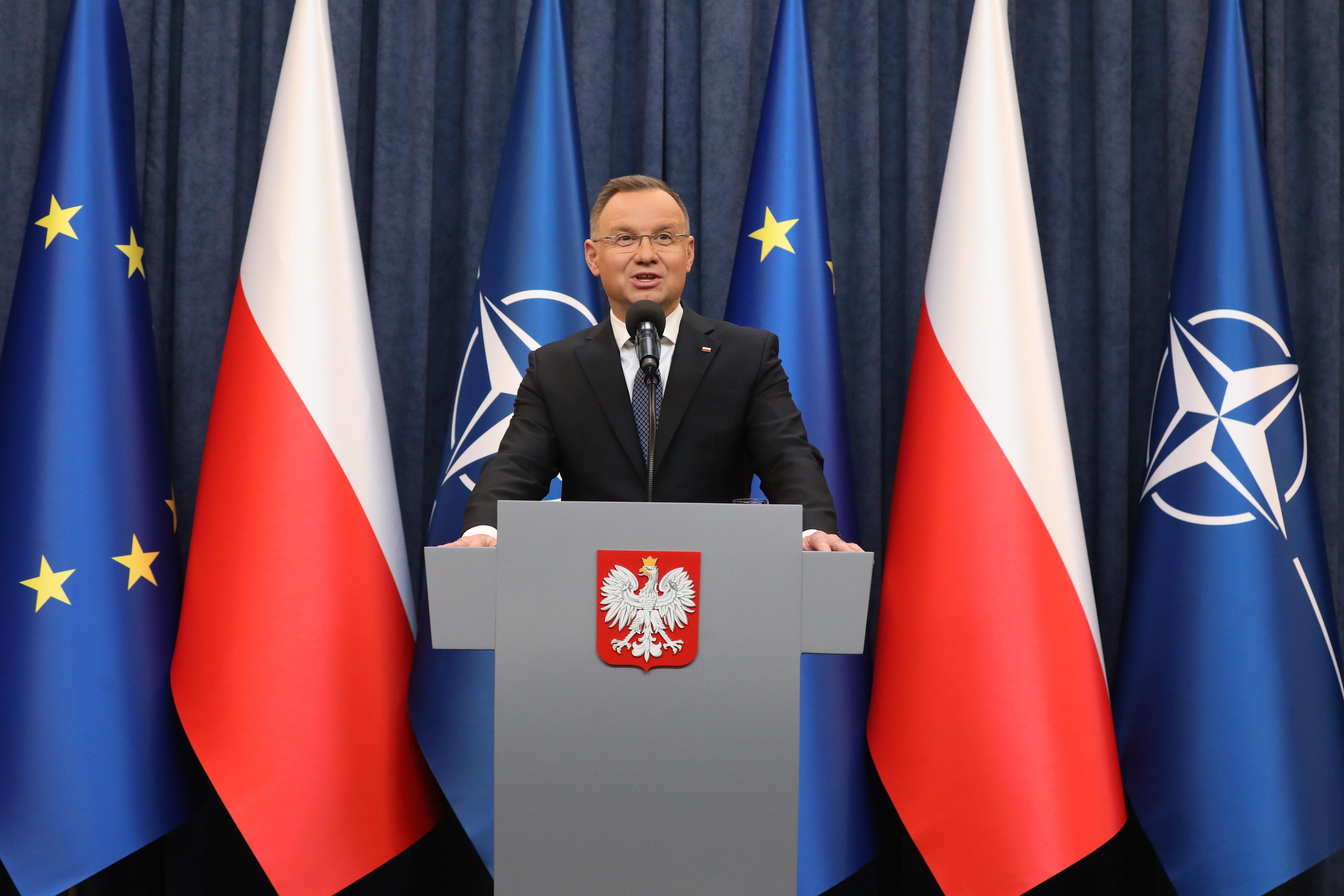 Prezydent Andrzej Duda przemawia zza mównicy z godłem Polski, za nim flagi Polski, Unii Europejskiej i NATO