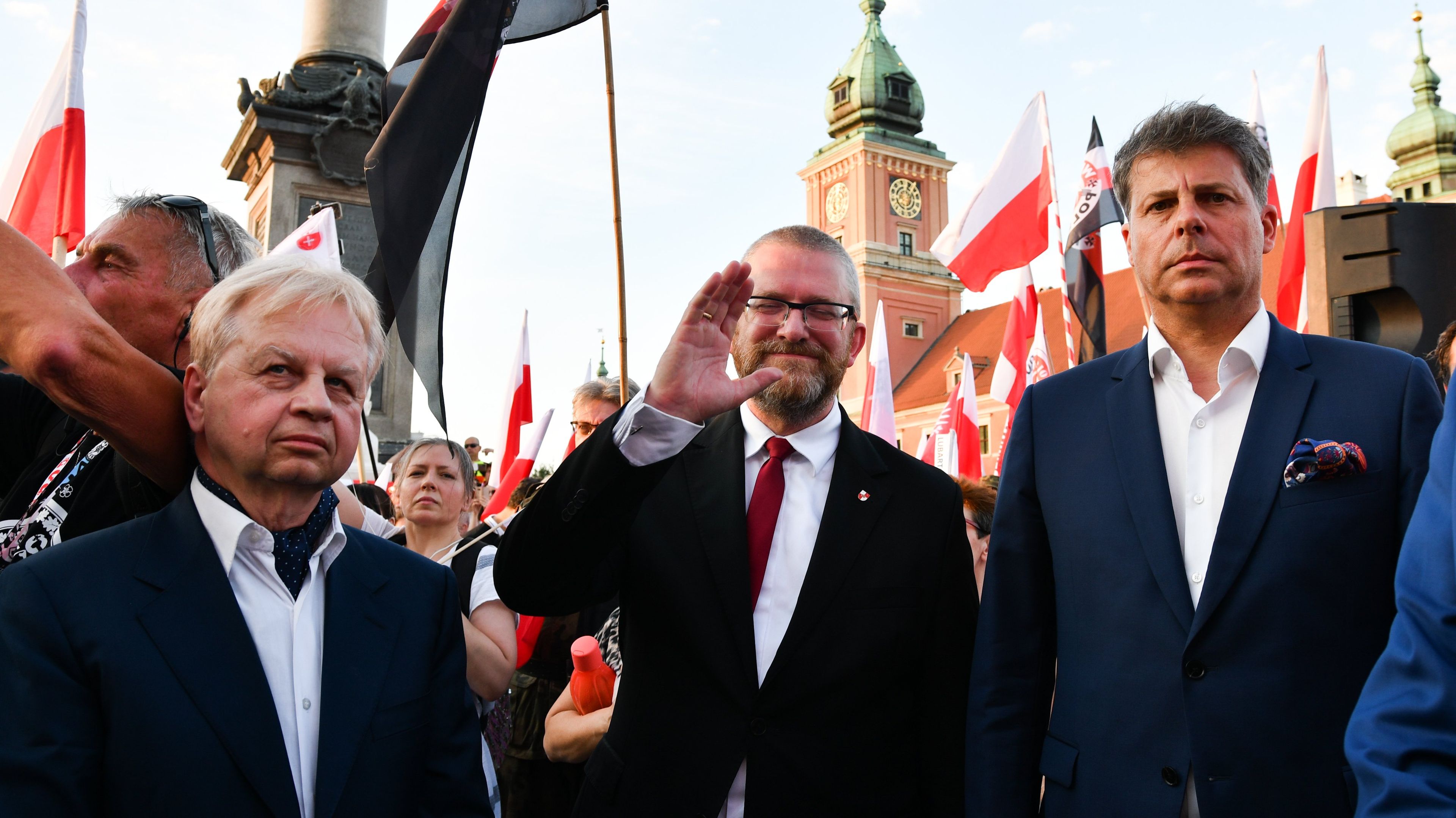 Grzegorz Braun i dwóch innych mężczyzn pod kolumną Zygmunta, w tle flagi