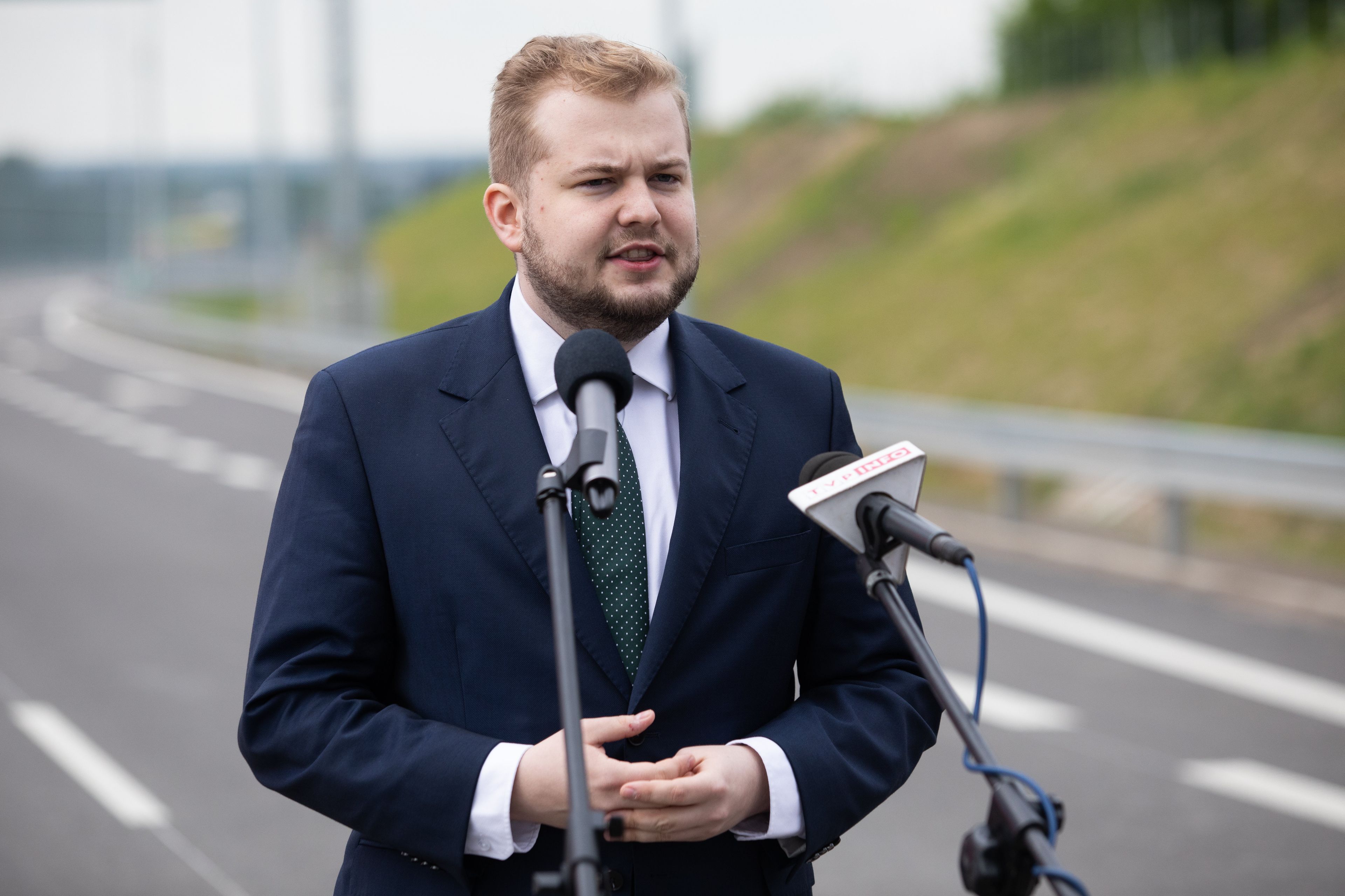 Michał Moskal w garniturze stoi przy mikrofonach na środku drogi S19