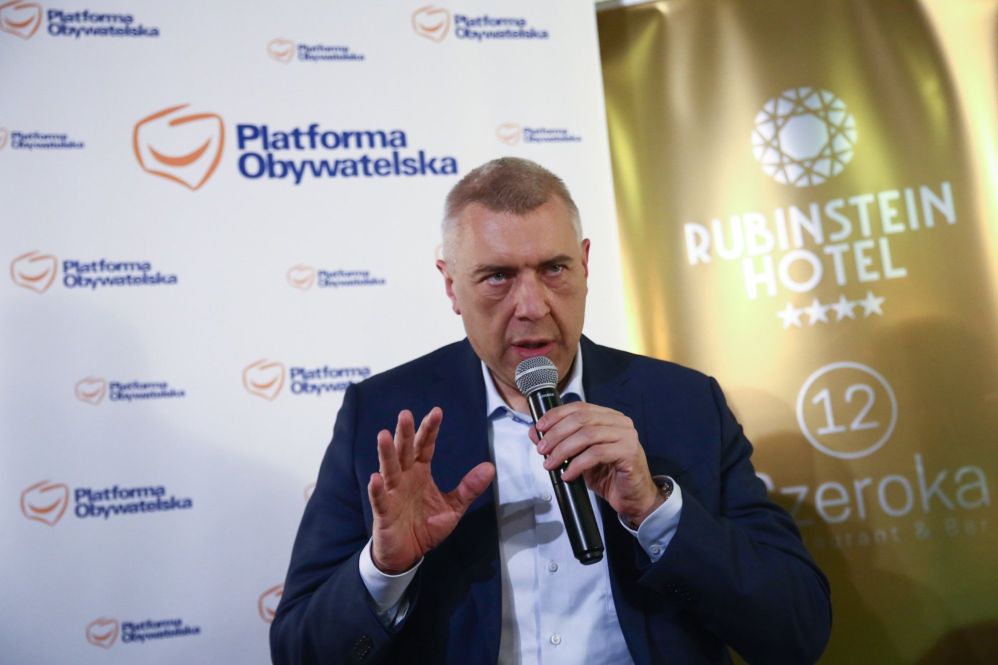 Roman Giertych na konferencji prasowej. Przemawia do mikrofonu, który trzyma w ręce. Ubrany w ciemną marynarkę i rozpiętą pod szyją jasną koszulę.