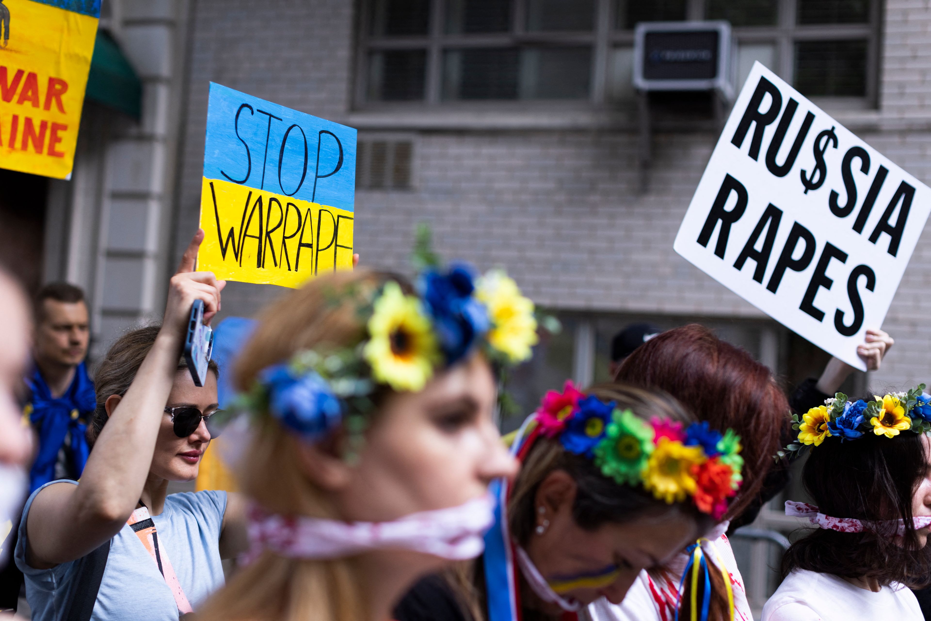 Demonstracja przeciwko rosyjskiej agresji na Ukrainę, na pierwszym planie dzieczyna w niebiesko-żółtym wianku na głowie, w tle plakat Russia Rapes - Rosja gwałci. Przemoc seksualna na wojnie