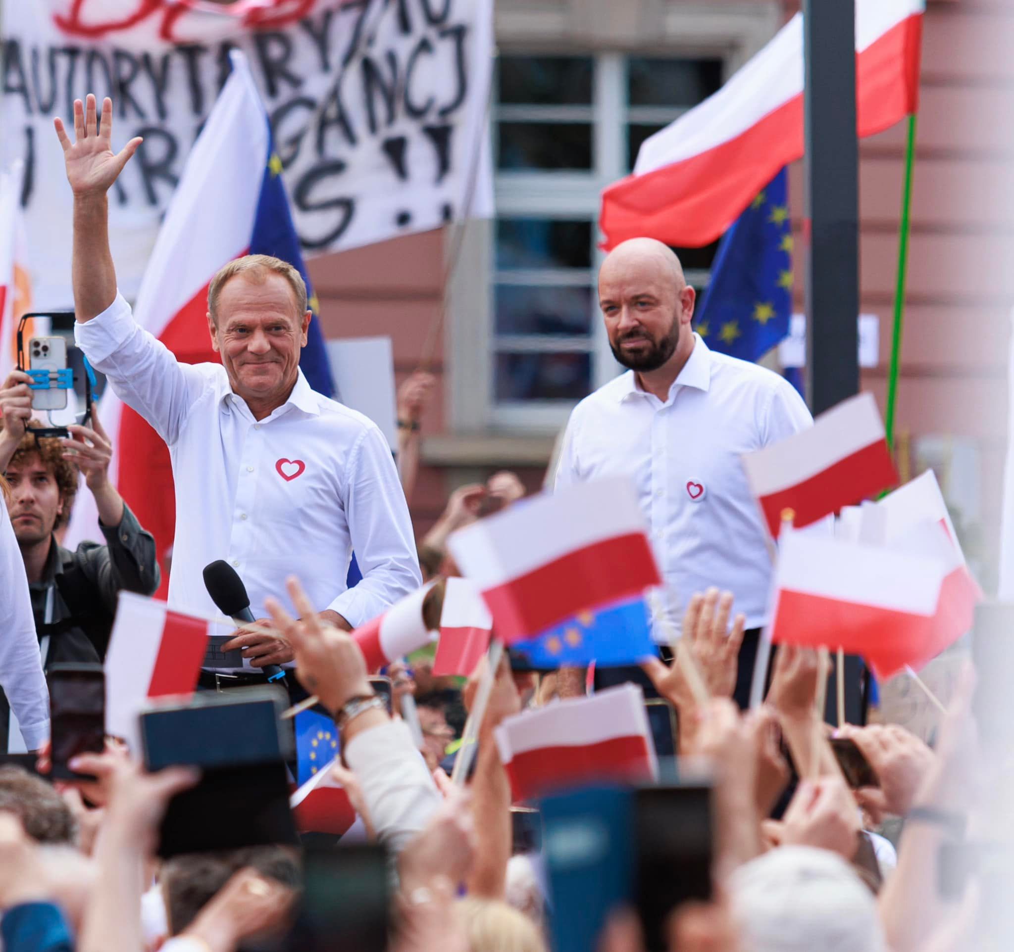 Dwóch mężczyzn w białych koszulach z małymi naklejkami czerwonych serduszek na piersi macha i uśmiecha się do tłumu wyborców machających biało-czerwonymi flagami