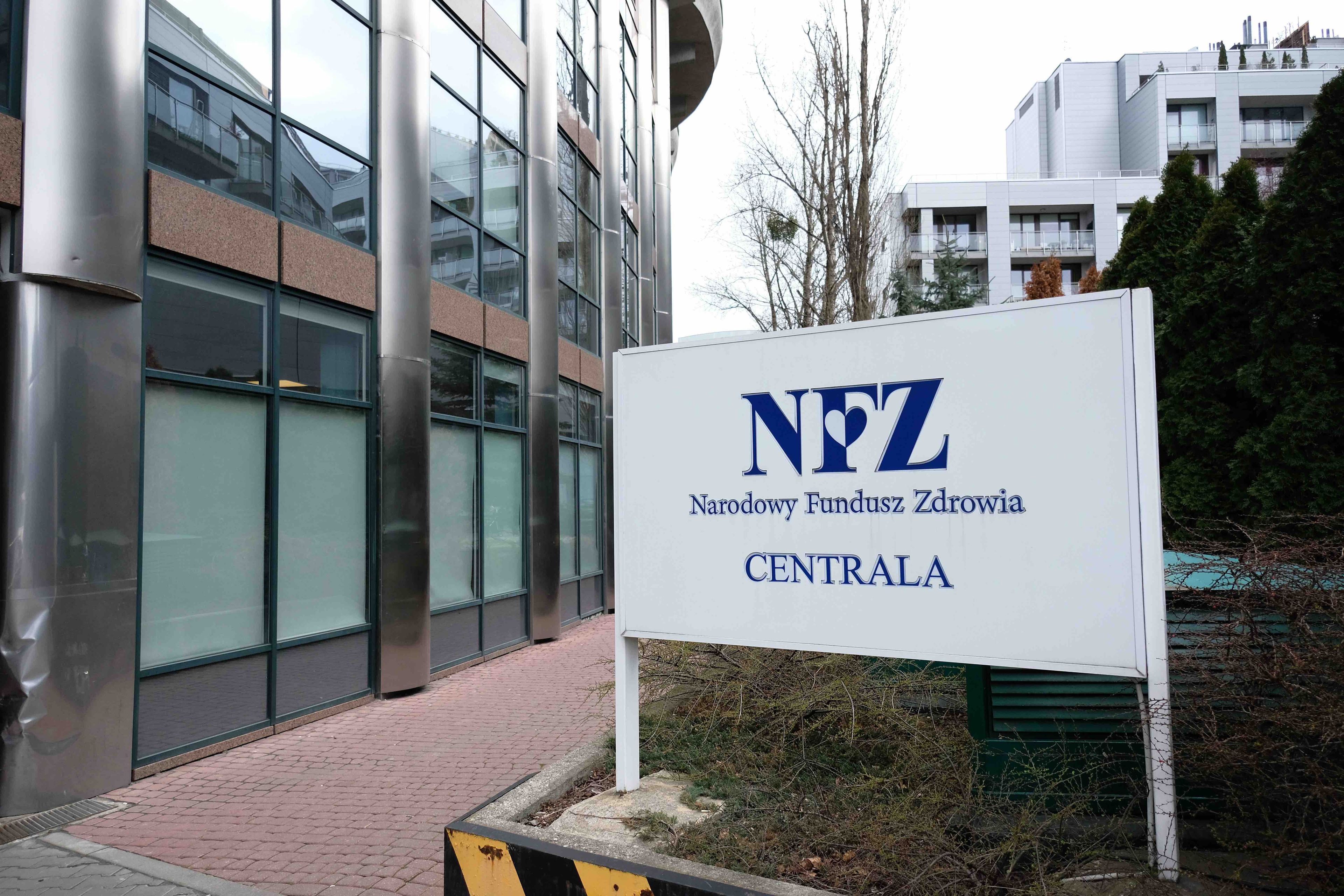 Fasada nowoczesnego biurowca, przed nią tablica z napisem Narodowy Fundusz Zdrowia Centrala