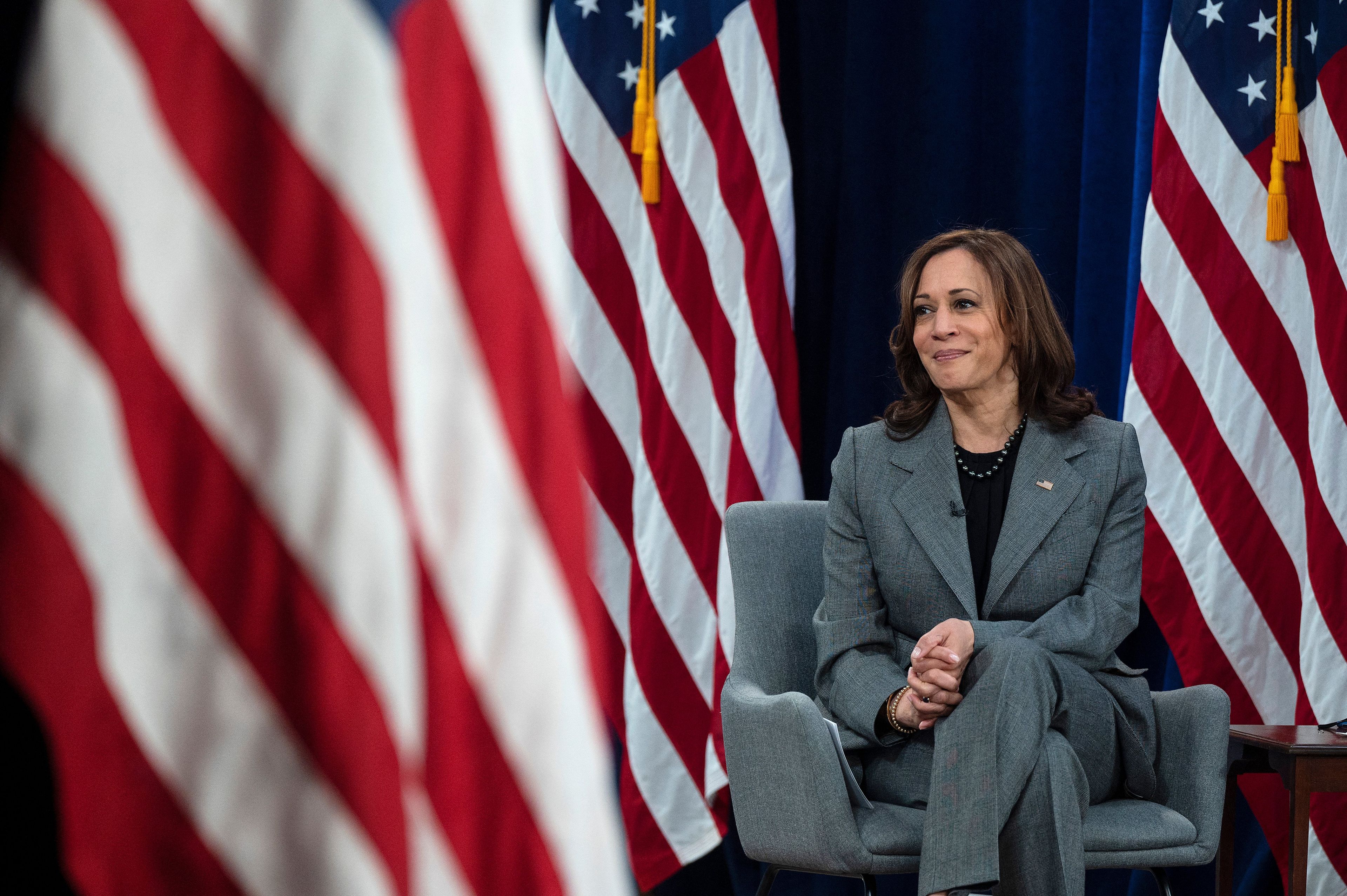 Kamala Harris ubrana w szaro-stalowy żakiet siedzi na krześle. Za nią flagi USA