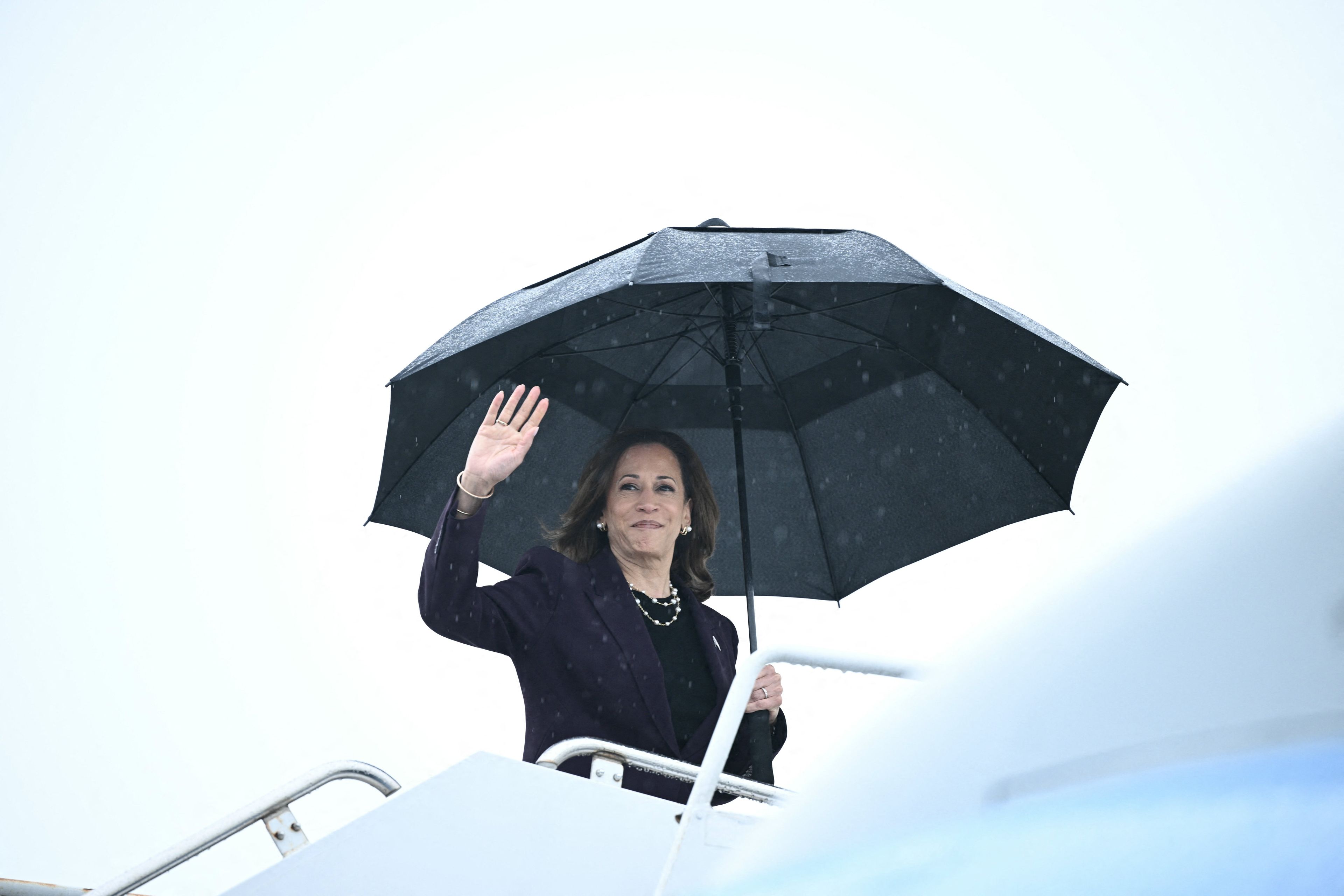 Kamala Harris wchodzi po schodkach do samolotu. W ręku trzyma rozłożony nad głową duży czarny parasol