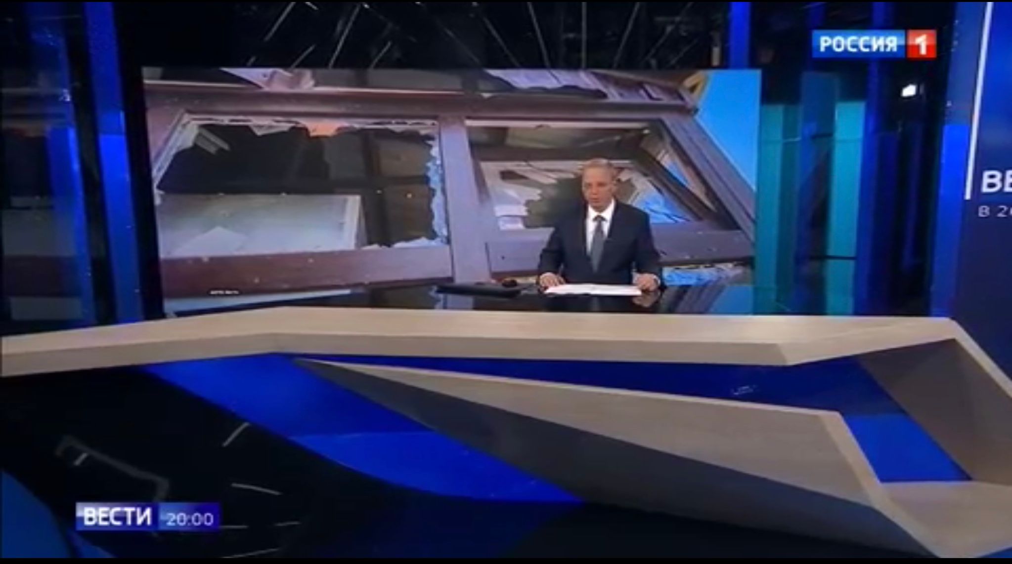 Studio telewizyjne. Za reporterem zdjęcie domu ze strzaskanymi szybami