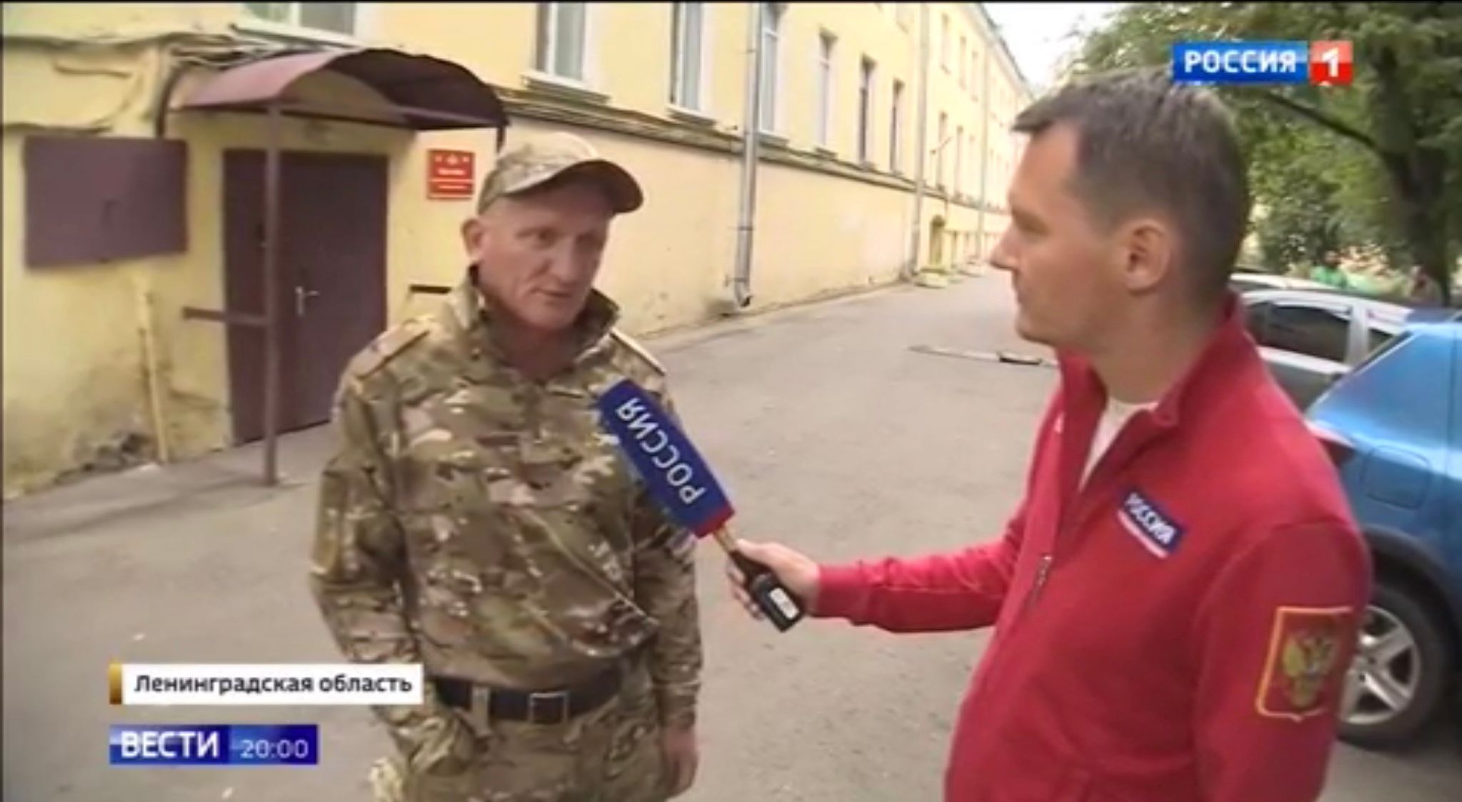 Męzczyzna w średnim wieku w mundurze i rosyjski reporter telewizyjny (ma mikrofon z napisem Rossija1)