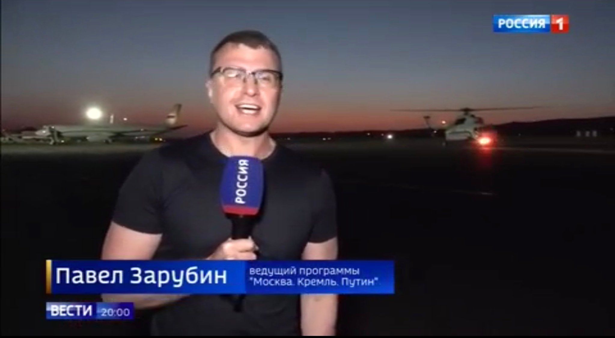 Rozentuzjazmowany mężczyzna mówi do mikrofonu z napisem "Rossija1", w tle helikopter lądujący o zmroku