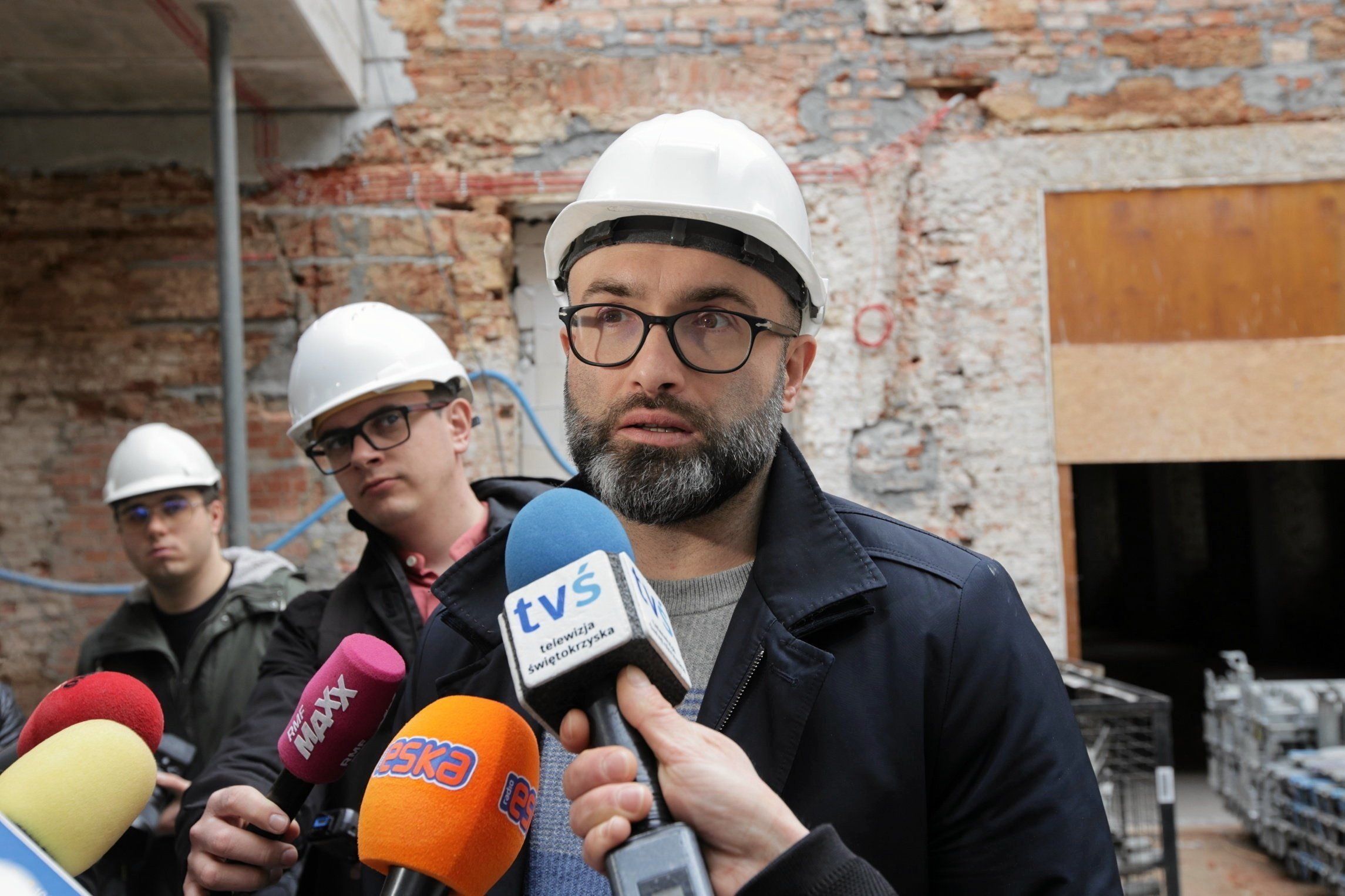 Mężczyzna w kasku budowlanym stoi na terenie budowy i udziela wywiadu dziennikarzom, za nim stoją dwaj inni mężczyźni w kaskach, w tle mur budynku. Michał Kotański