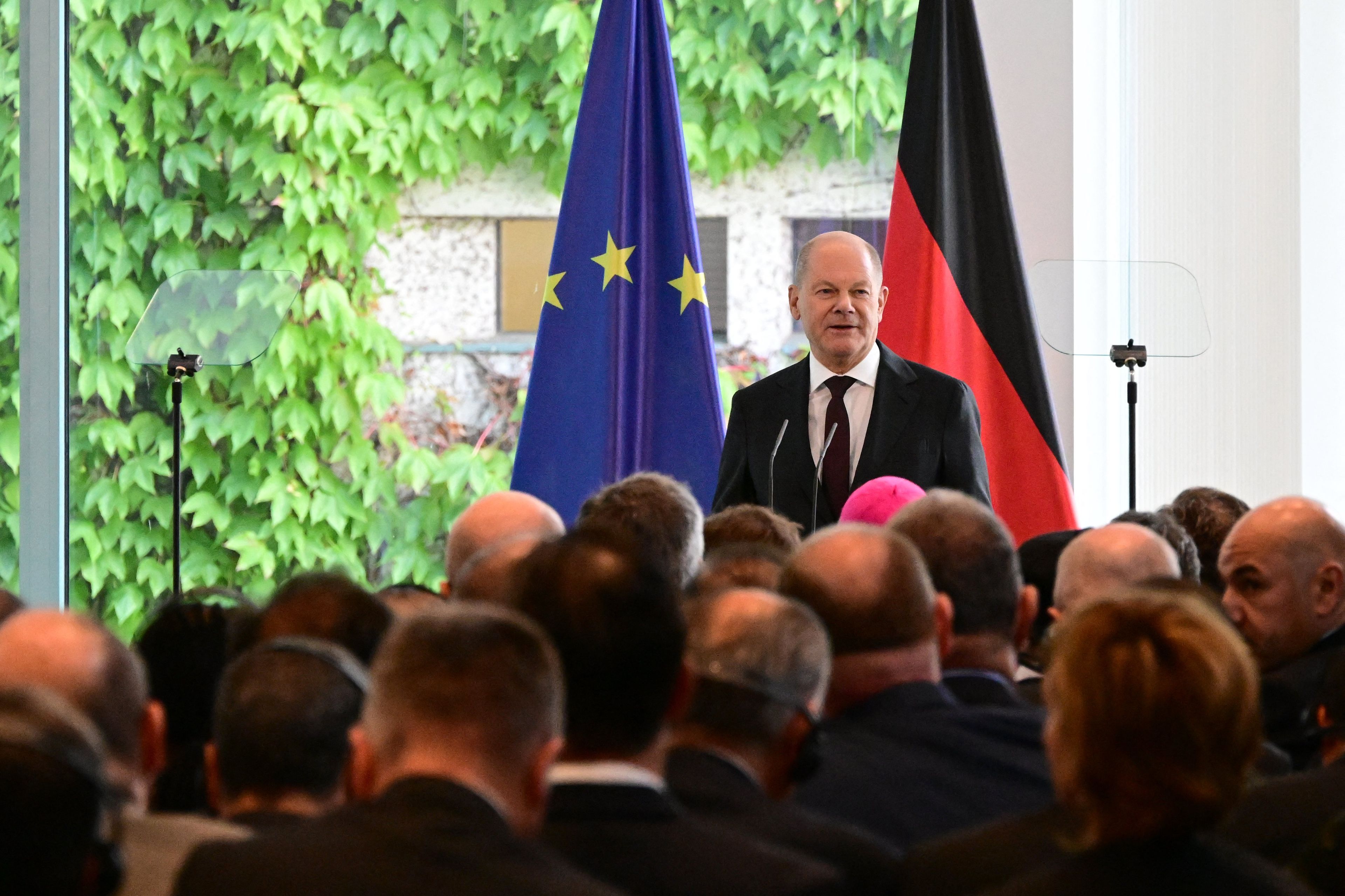 Łysięjący mężczyzna w garniturze stoi na tle flag Niemiec i Unii Europejskiej