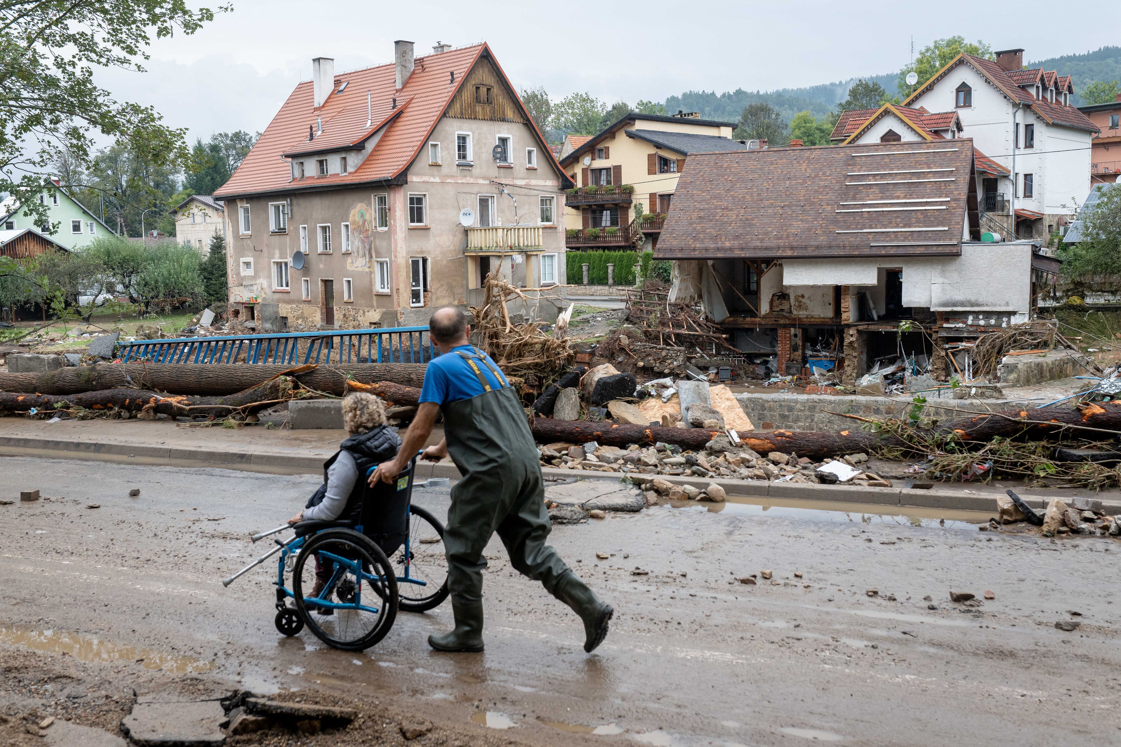 Mężczyzna pcha kobietę na wózku dla osób niepełnosprawnych. Wokół miasto zniszczone przez powódź