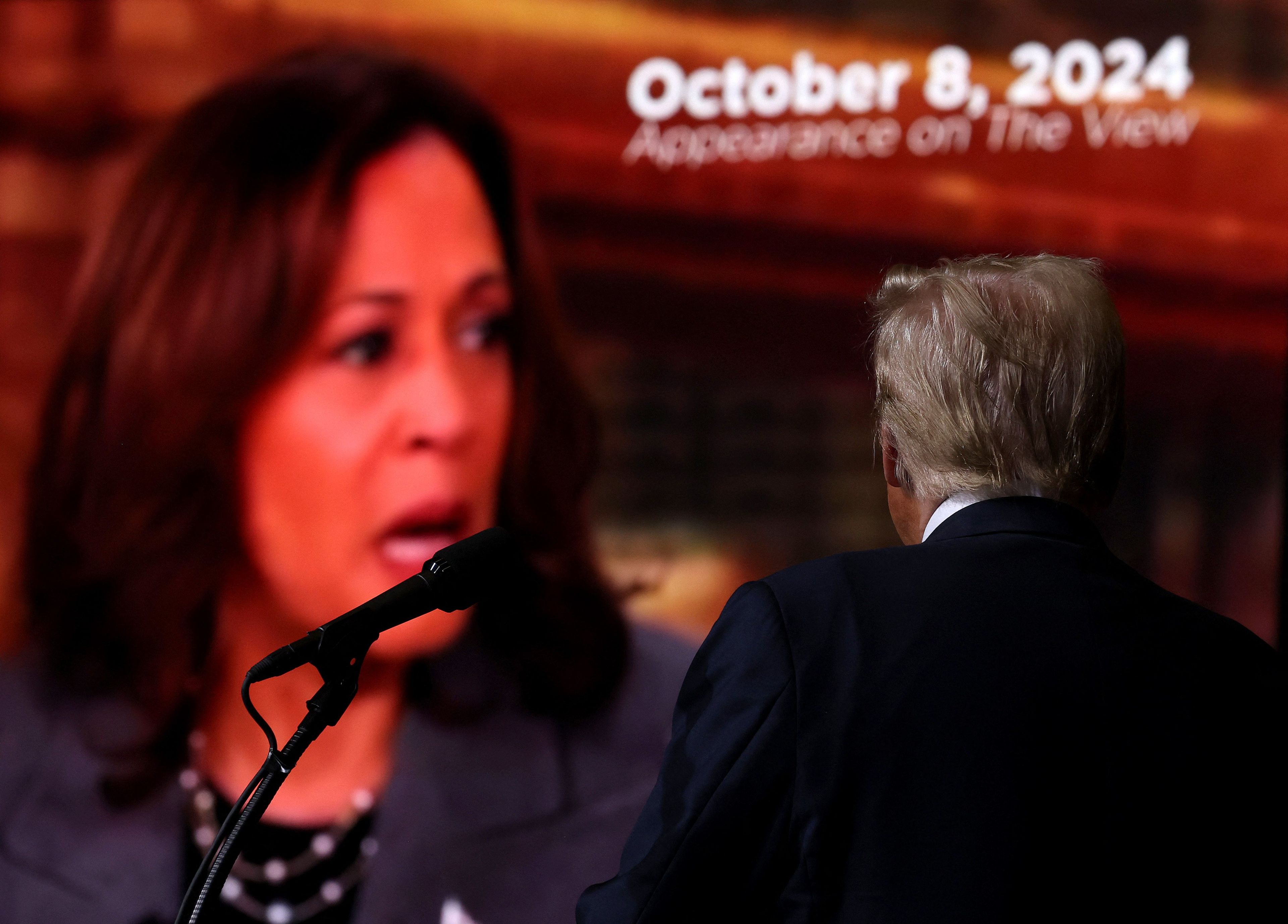 Donald Trump (ujęcie od tyłu) stoi przed wielkim ekranem, na którym widać twarz Kamali Harris