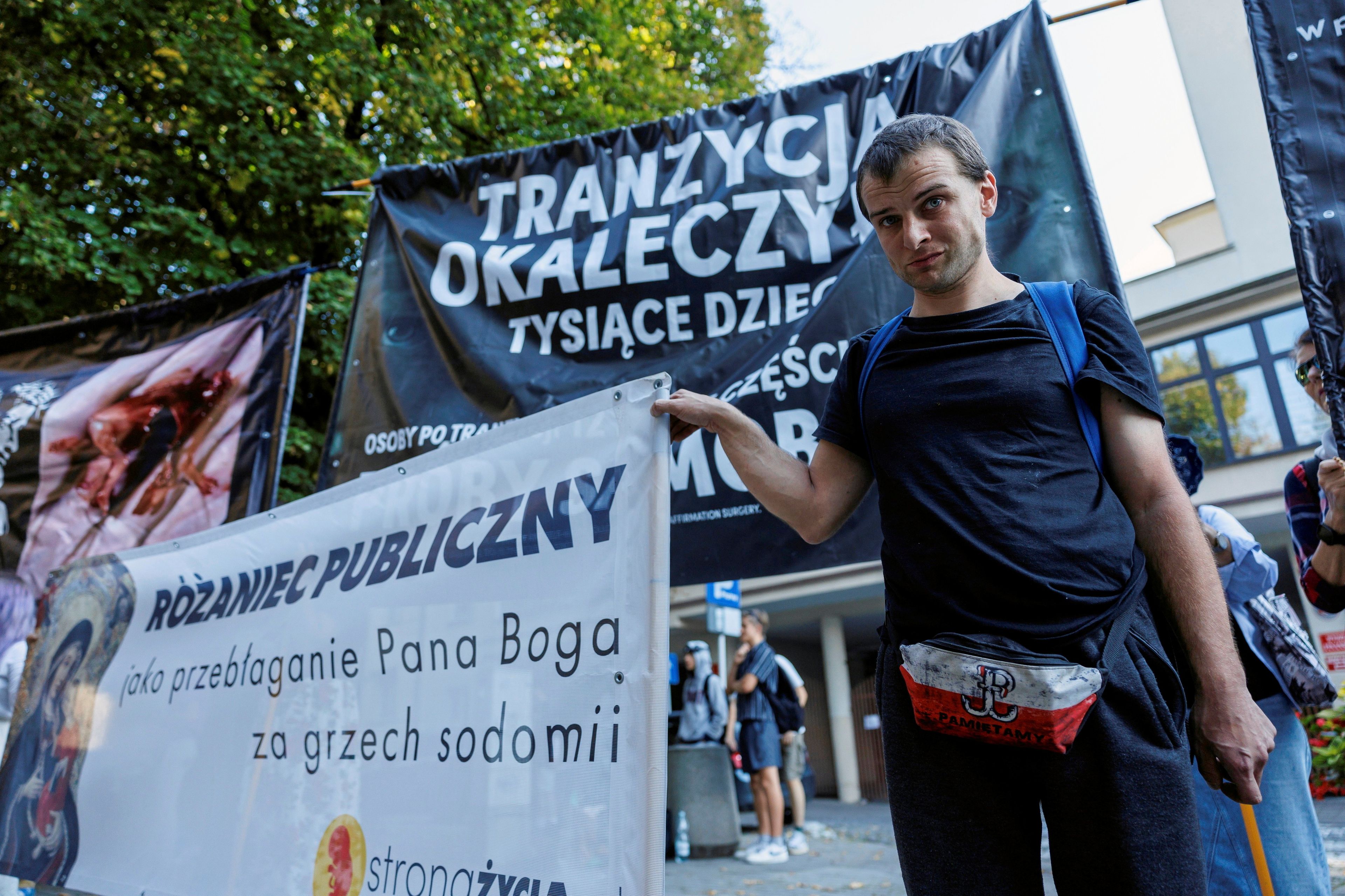 21.09.2024, Rzeszów. Kontrmanifestacja środowisk prawicowych na trasie V Marszu Równości w Rzeszowie. Młody mężczyzna ubrany w czarny t-shirt i biał-czerwoną nerkę ze znakiem Polski Walczącej trzyma transparent z napisem "Różaniec publiczny jak przebłaganie Pana Boga za grzech sodomii". Za nim transparent z napisem "Tranzycja okaleczyła tysiące dzieci"