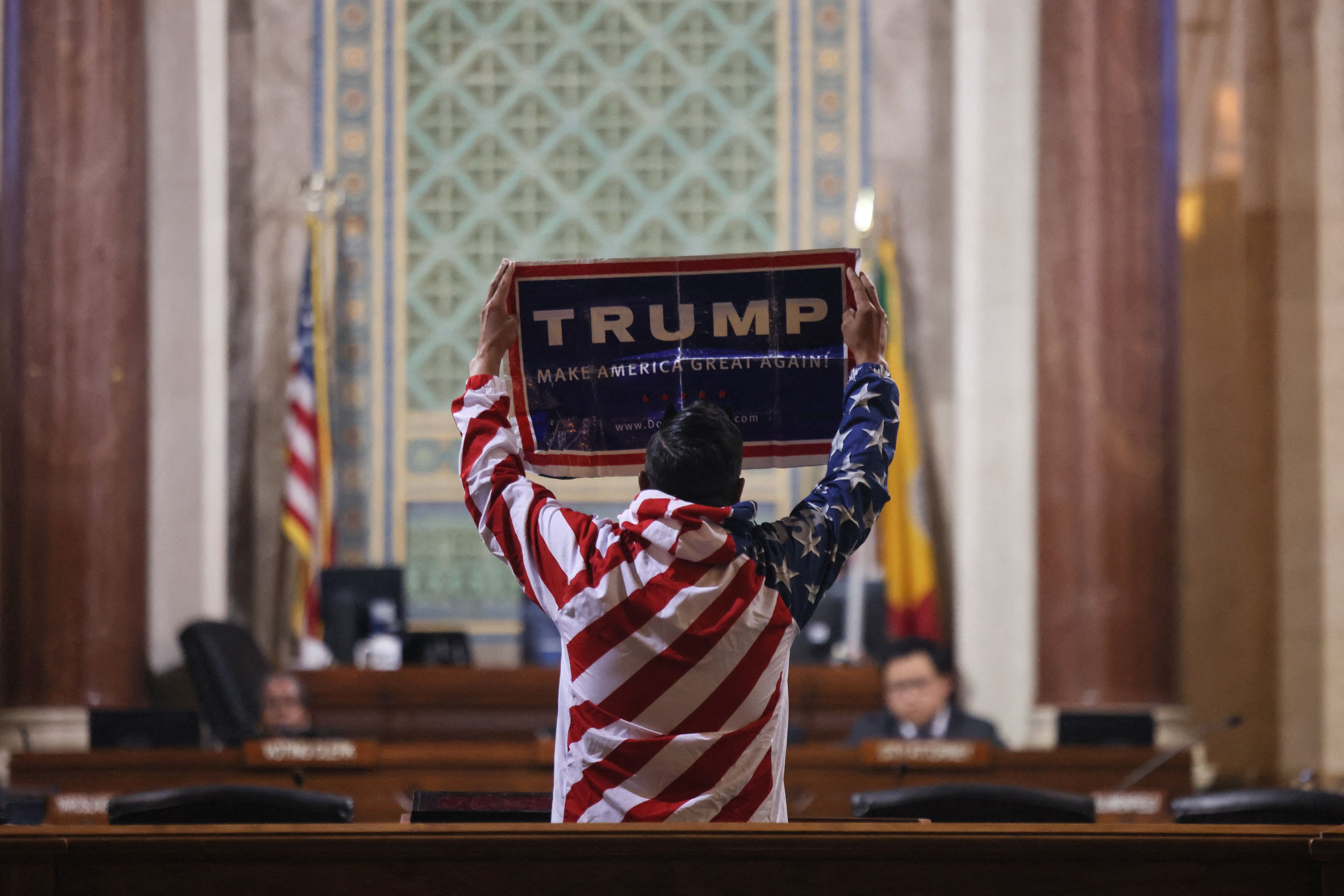 Mężczyzna w koszuli ze wzorem amerykańskiej flagi trzyma nad głową plakat: Trump. Make America Great Again