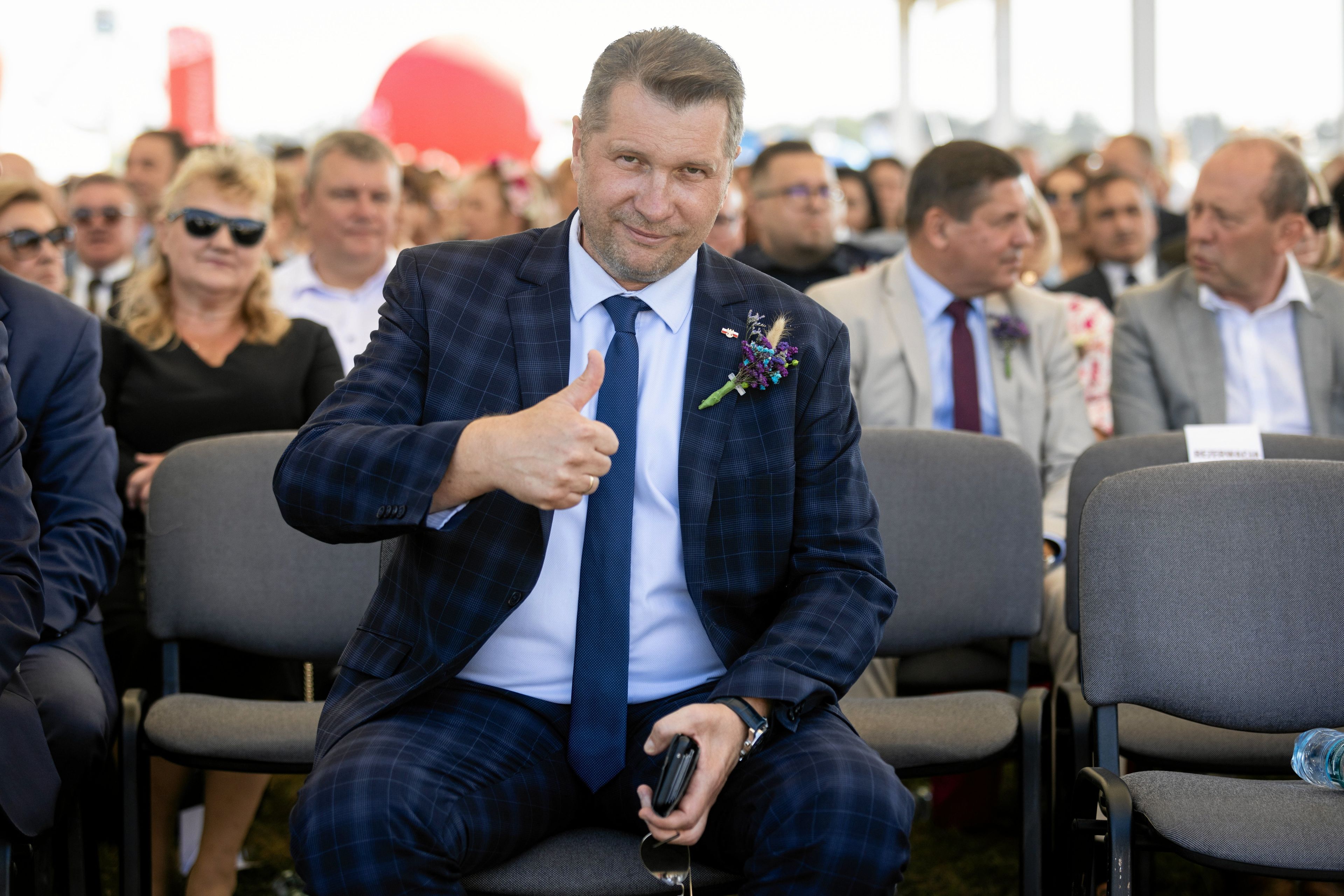 Mężczyzna w średnim wieku w kraciastej marynarce. Pokazuje uniesiony kciuk w stronę obiektywu