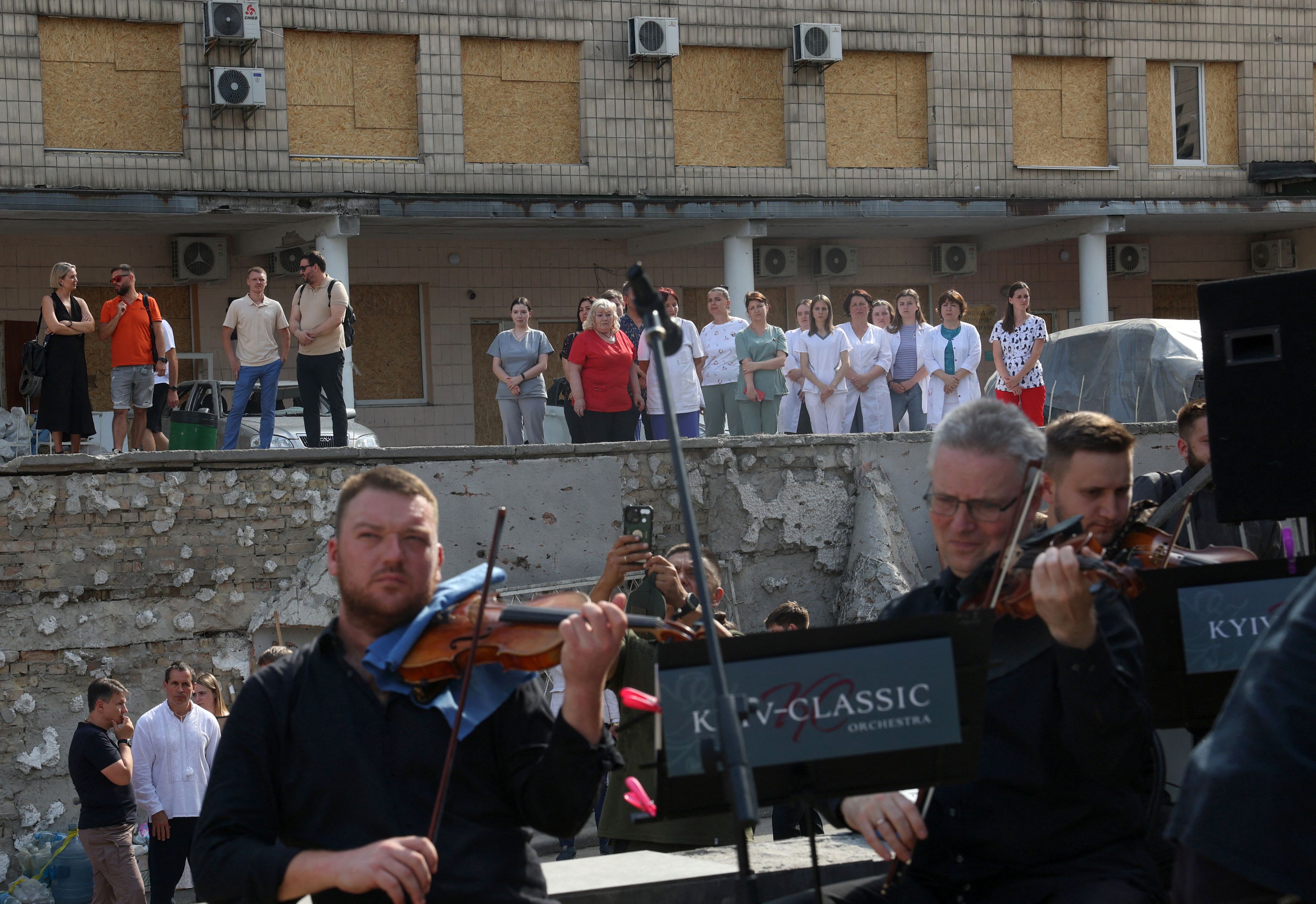 Na pierwszym planie - orkiestra, w tle - pracownicy medyczni w ruinach