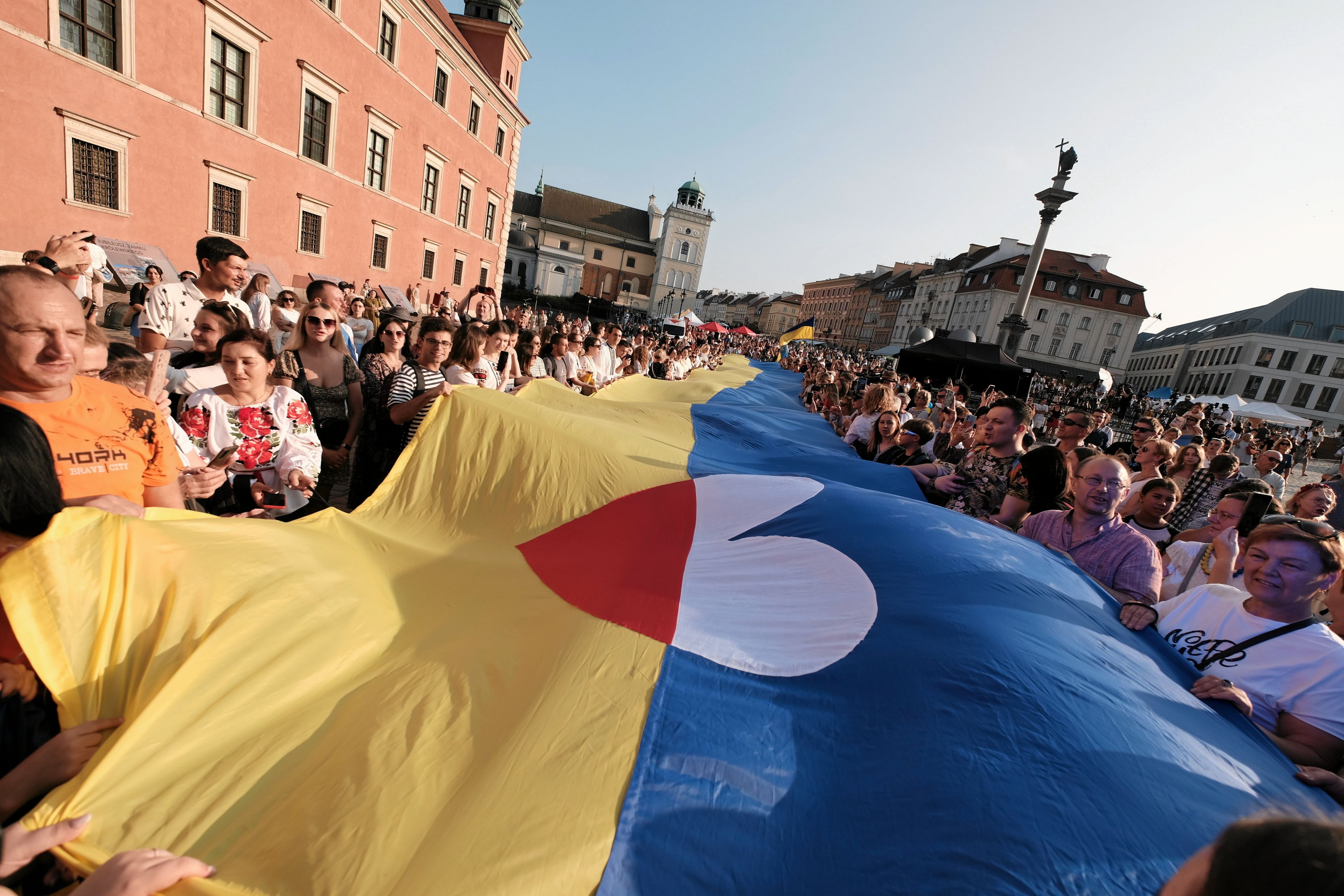 Na Placu Zamkowym w Warszawie tłum rozwija ukraińską flagę. Na środku jest serce w barwach polskiej flagi