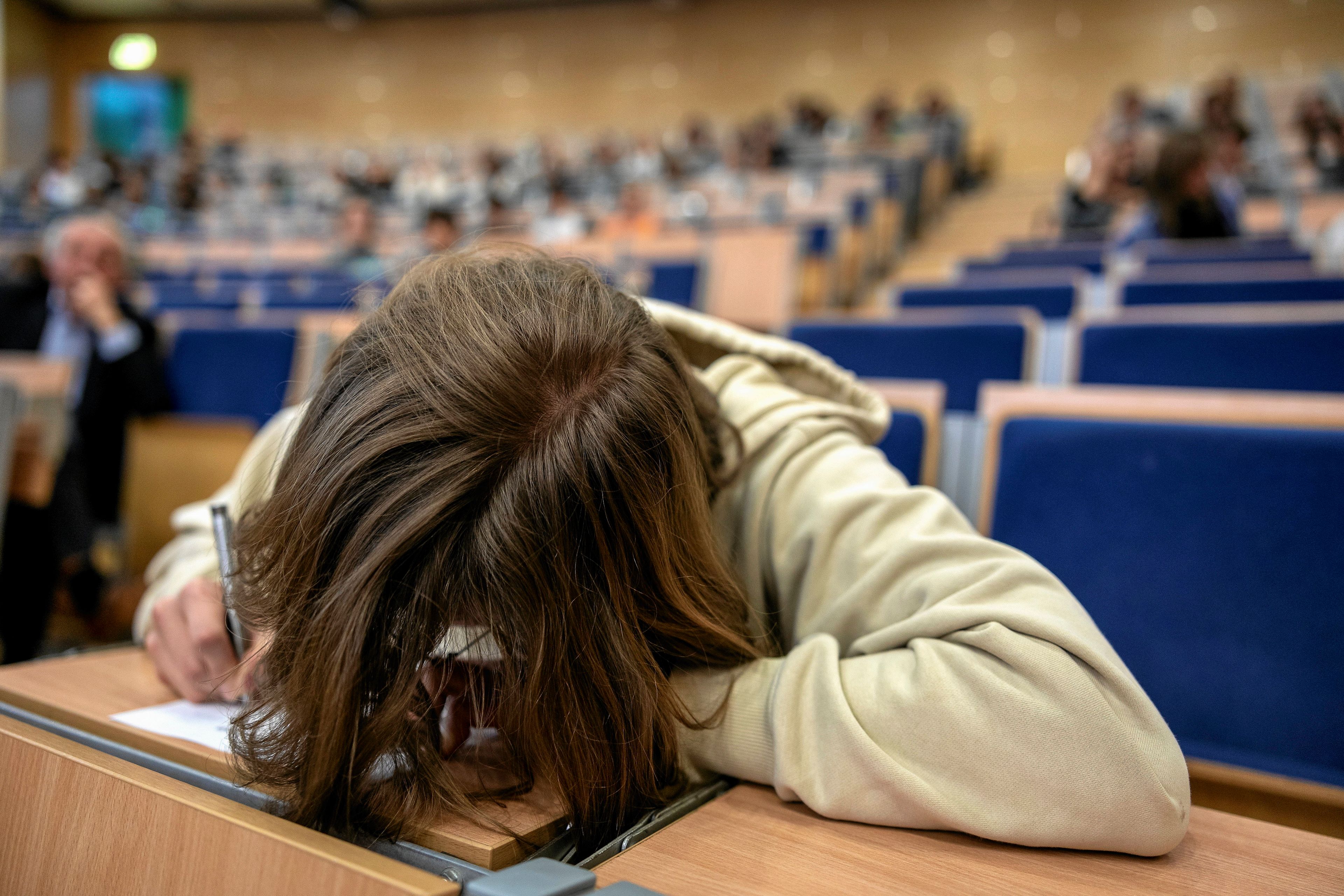 Młoda osoba z głową na biurku, z tyłu uniwersytecka aula