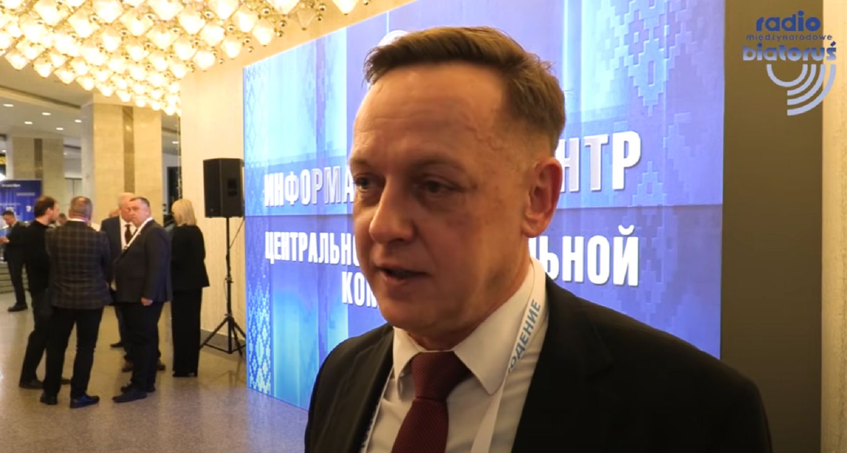 Tomasz Szmydt - mężczyzna z krótkimi włosami, w garniturze, białej koszuli i krawacie, stoi w holu, na tle niebieskiej ścianki z napisem