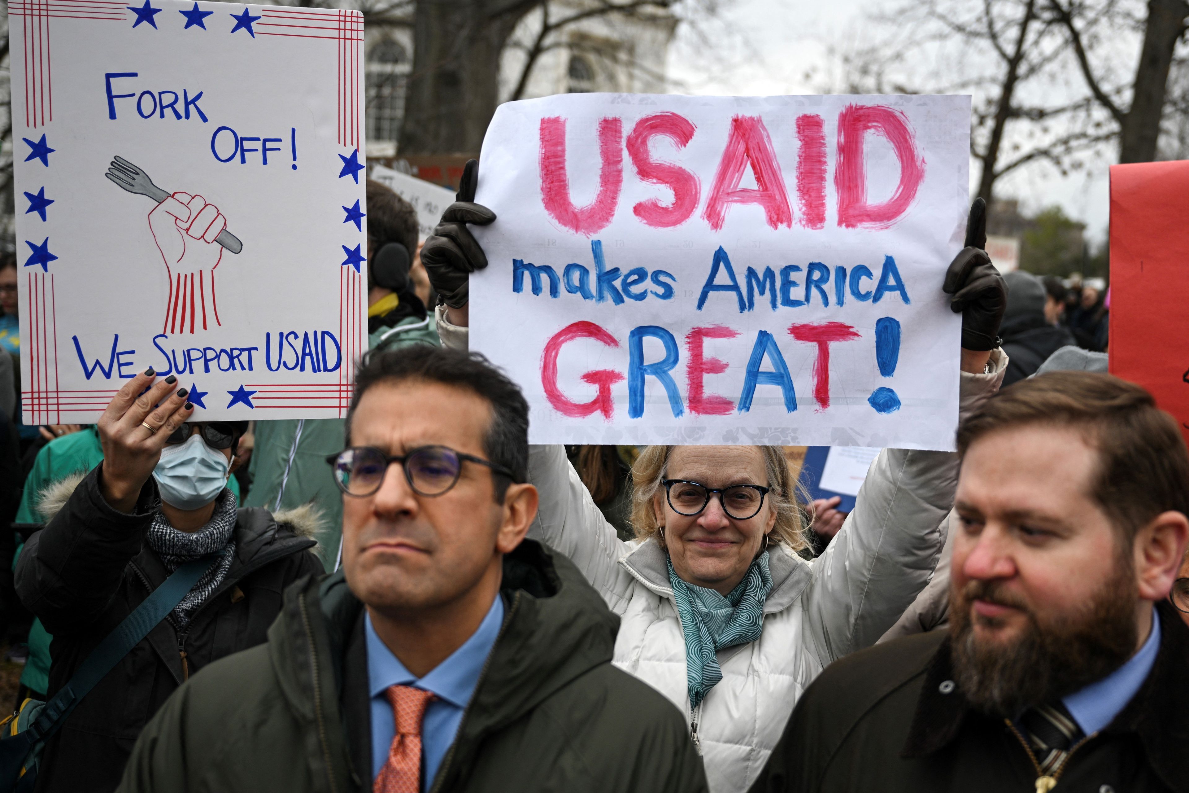 Tłum ludzi na demonstracji, jedna z kobiet trzyma nad głową biały transparent z napisem "USAID makes America GREAT!"