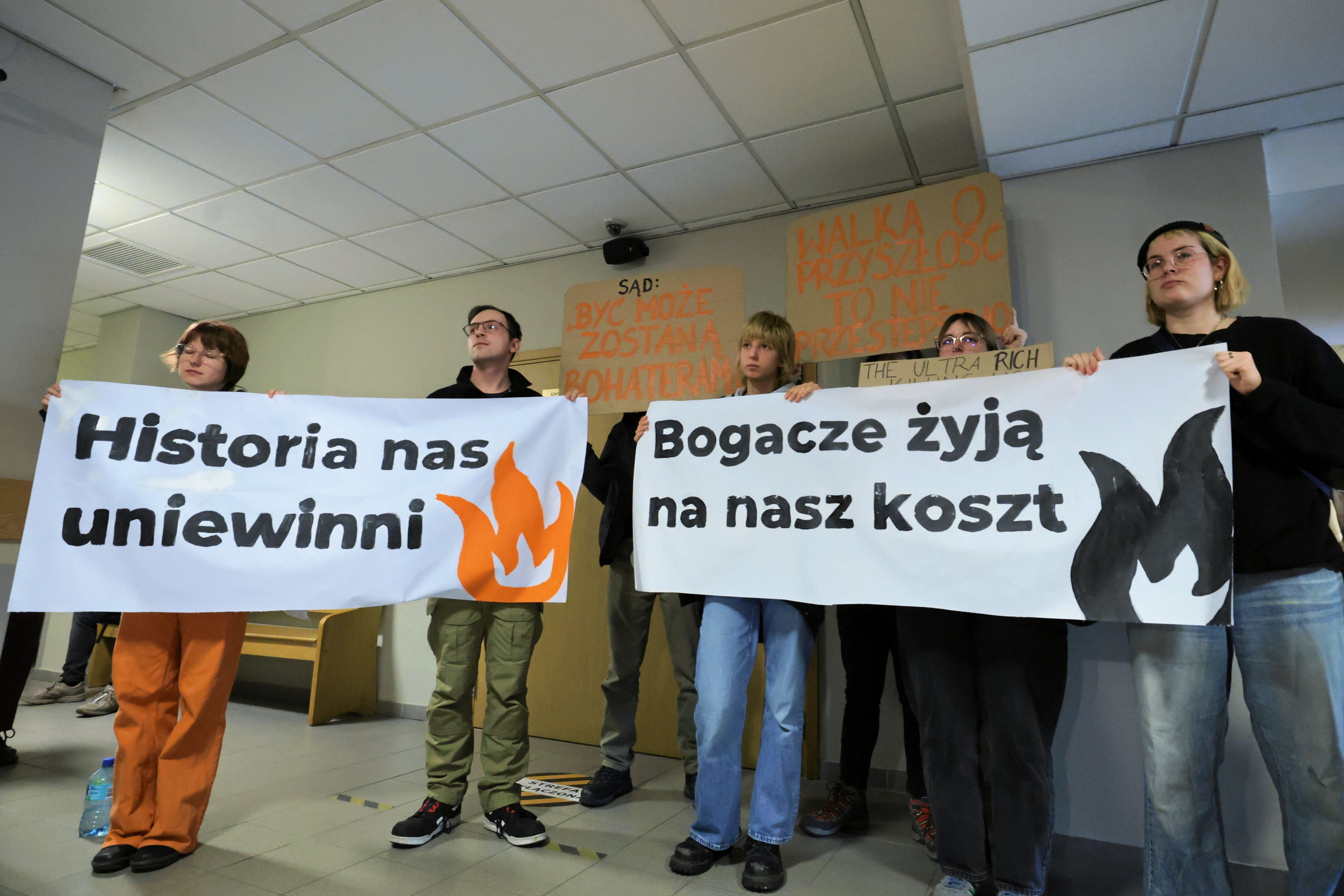 Młodzi ludzie stoją w sądzie z transparentami "Historia nas uniewinni" i "Bogacze żyją na nasz koszt"