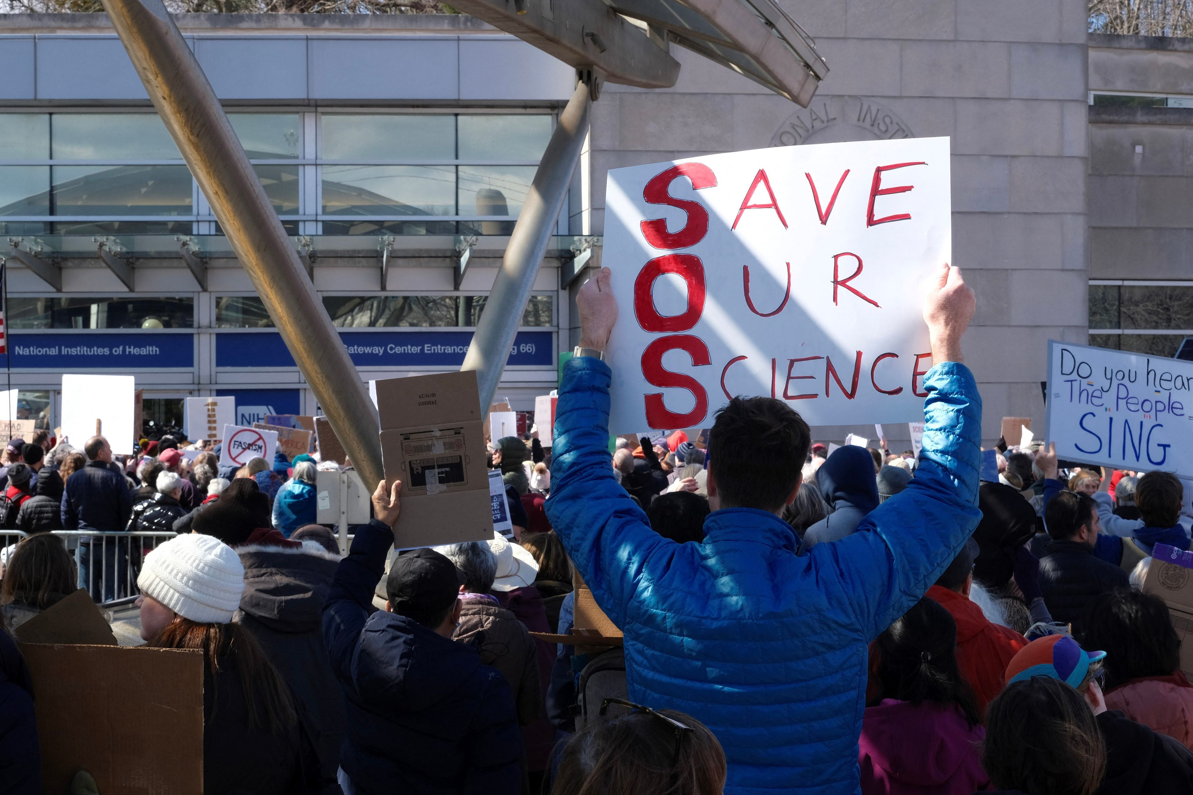 Tłum demosntrantów, jeden na pierwszym planie trzyma baner z napisem SOS, Save our Science