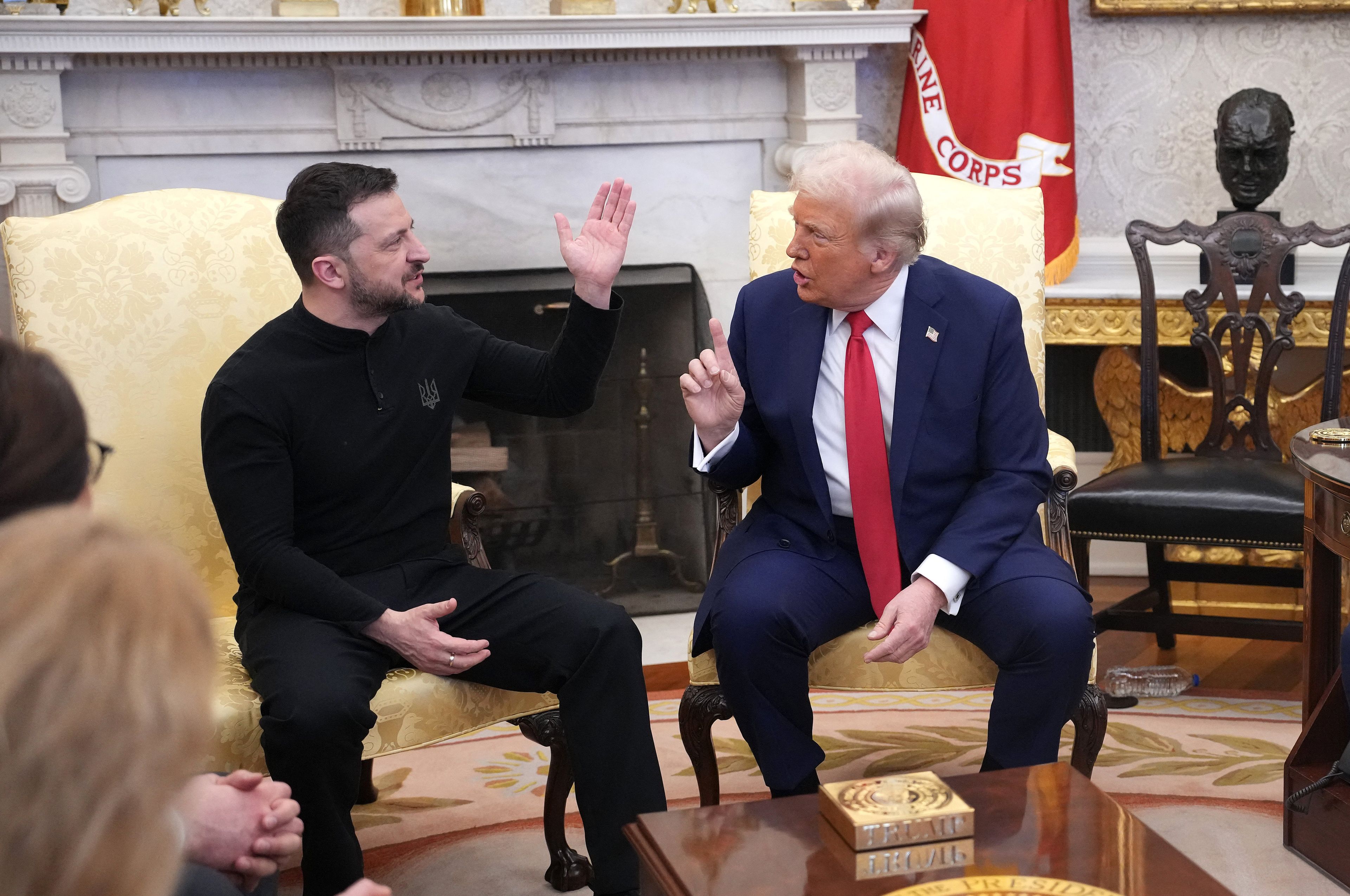 Mężczyzna ubrany na czarno, z czarną brodą, siedzi obok wyższego mężczyzny w garniturze i czerwonym krawacie z żółtymi włosami, obaj gestykulują.