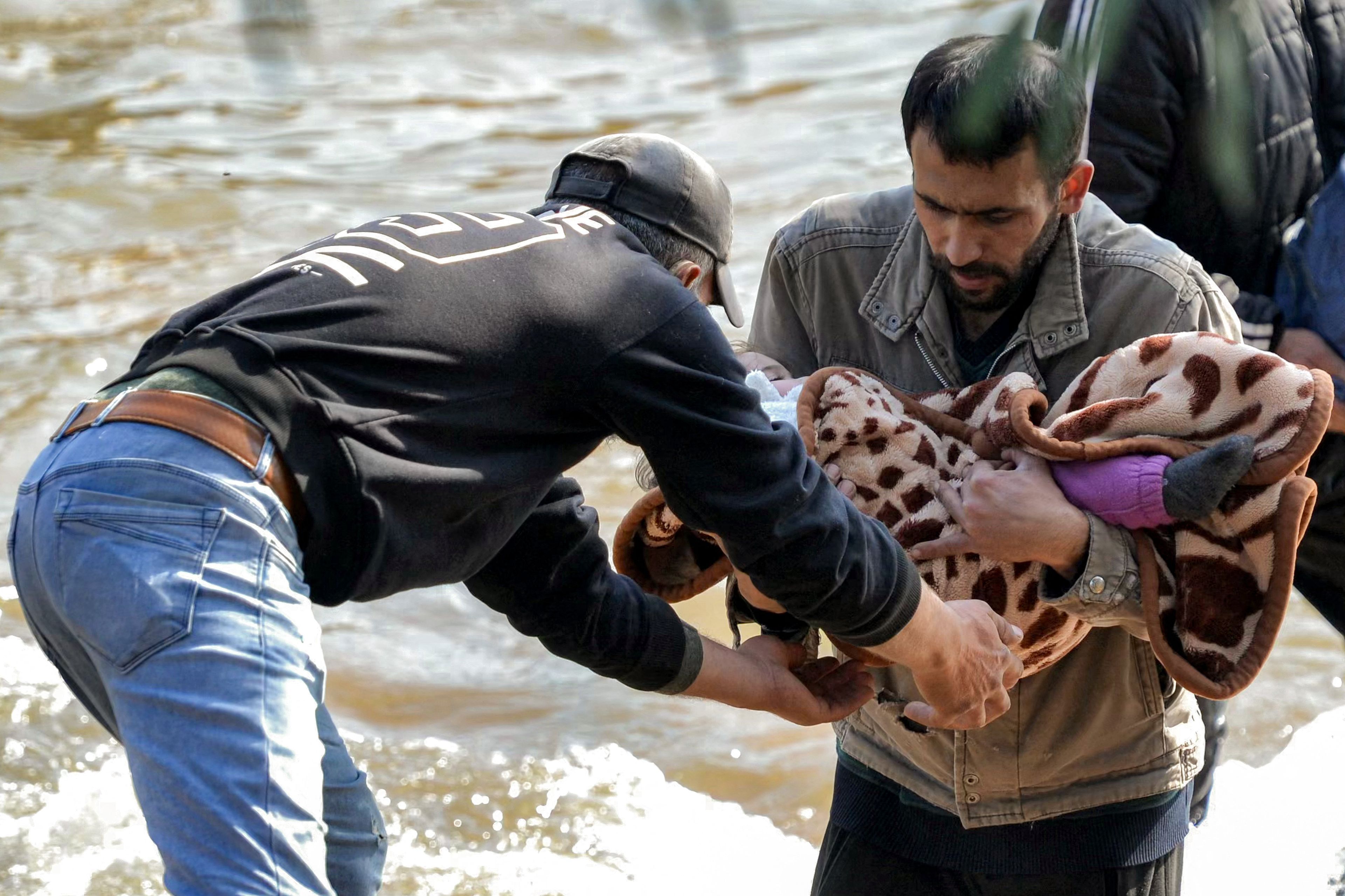 A man receives a blanketed child from another crossing the Nahr al-Kabir river, forming the border between Syria's western Latakia province and northern Lebanon in the Hekr al-Daher area on March 11, 2025, as families from Syria's Alawite minority enter Lebanon to flee from sectarian violence in their heartland along Syria's Mediterranean coast. (Photo by Fathi AL-MASRI / AFP)