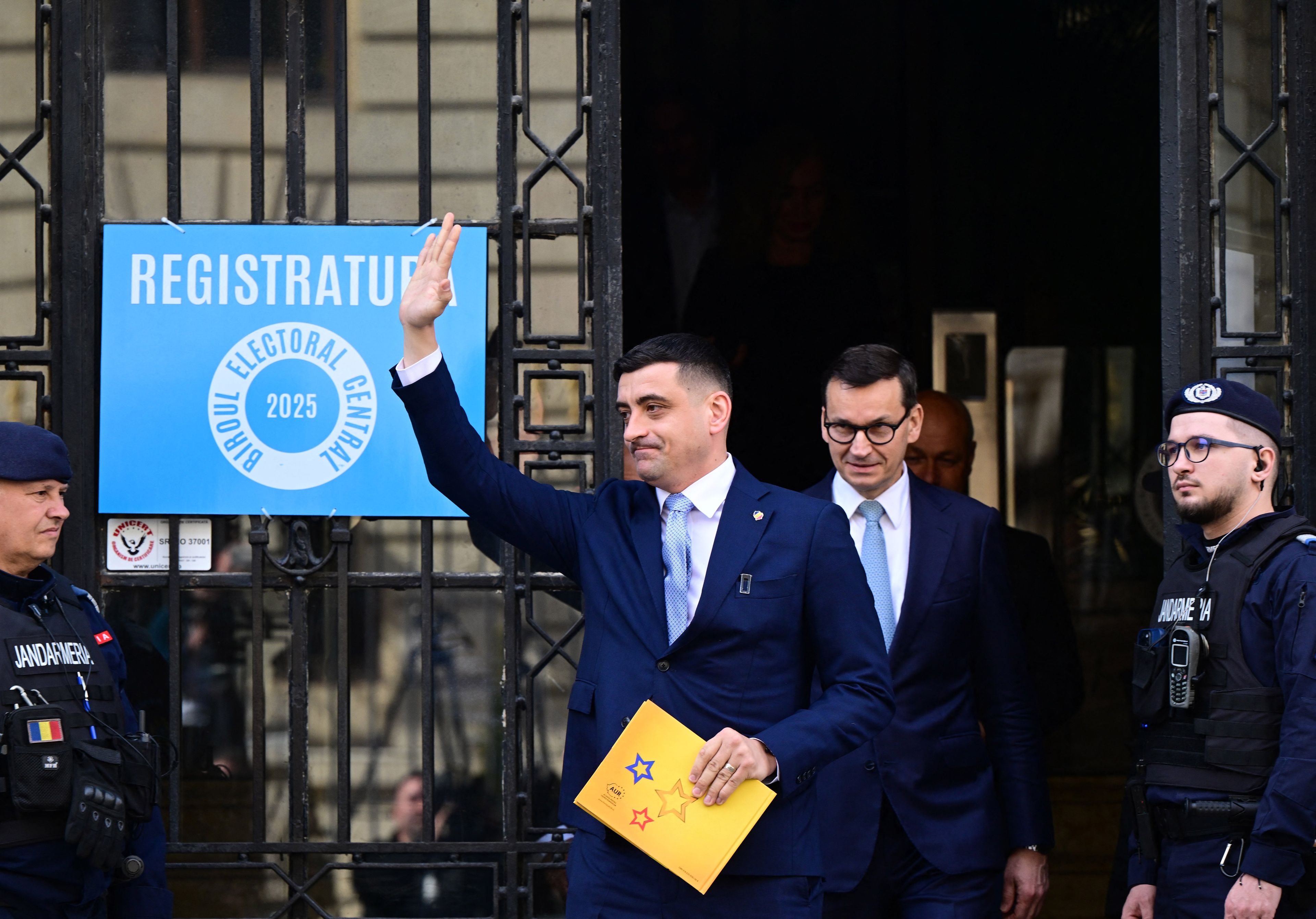 Dwóch mężczyzn wychodzi z siedziby komisji wyborczej, ten z porzudu podnosi rękę , ten z tyłu, Morawiecki, uśmiecha się