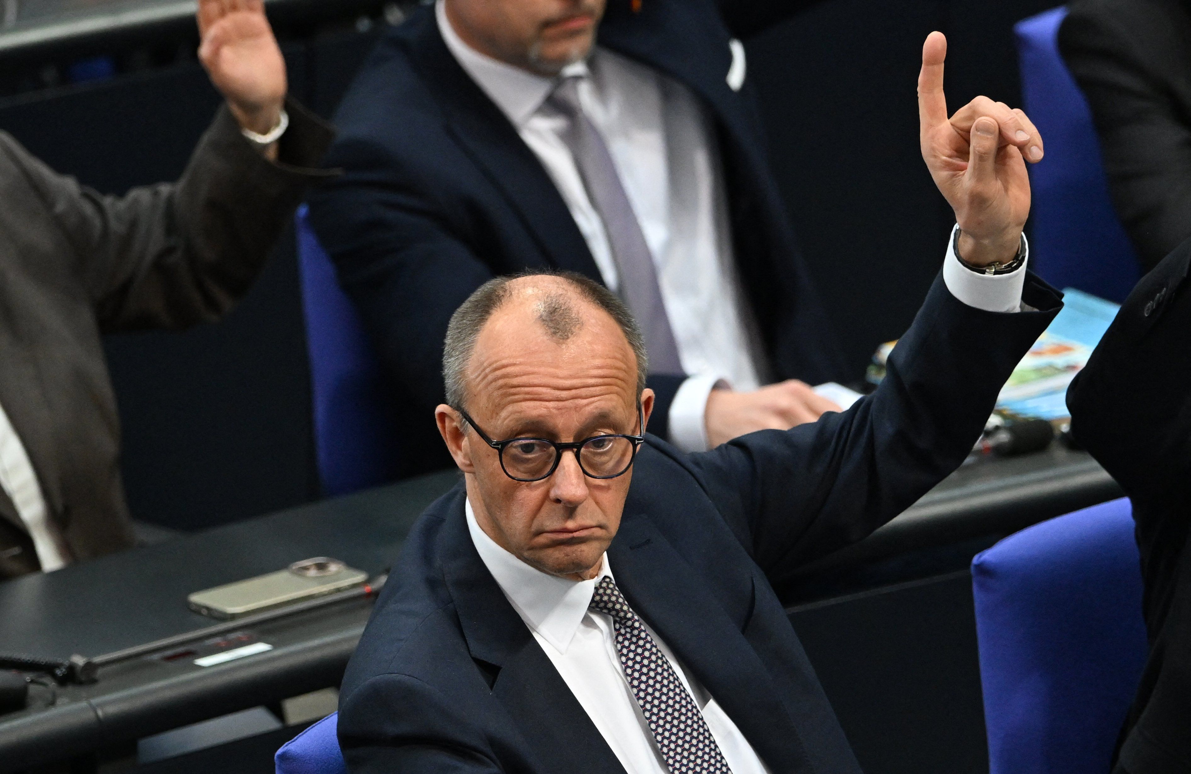 Friedrich Merz, mężczyzna w średnim wieku z łysiejącą głową i w okularach siedzi w Bundestagu i podnosi rękę w trakcie głosowania