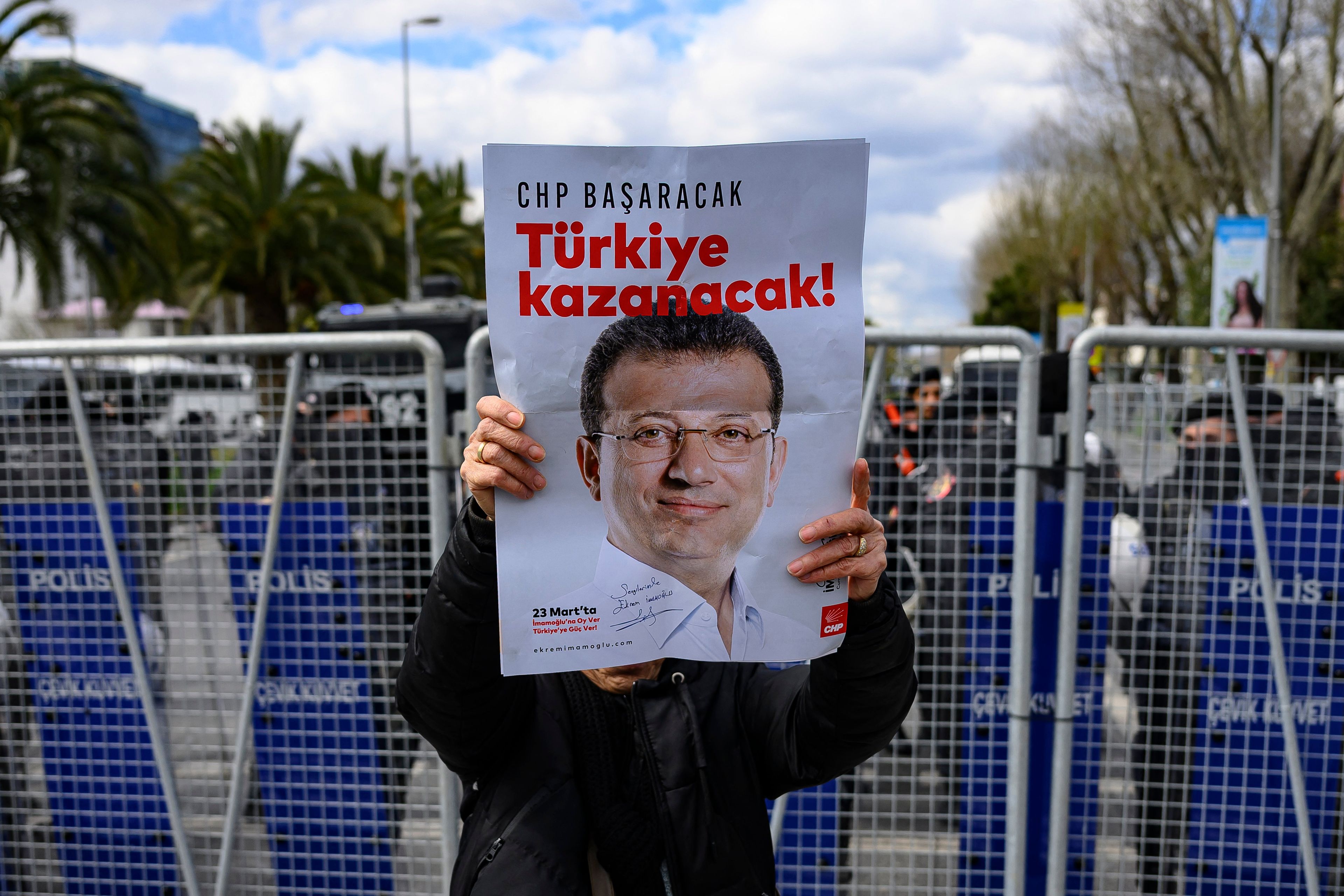 Protestujący trzyma plakat z wizerunkiem Ekrema Imamoglu, zatrzymanego burmistrza Stambułu, w tle blokada policyjna