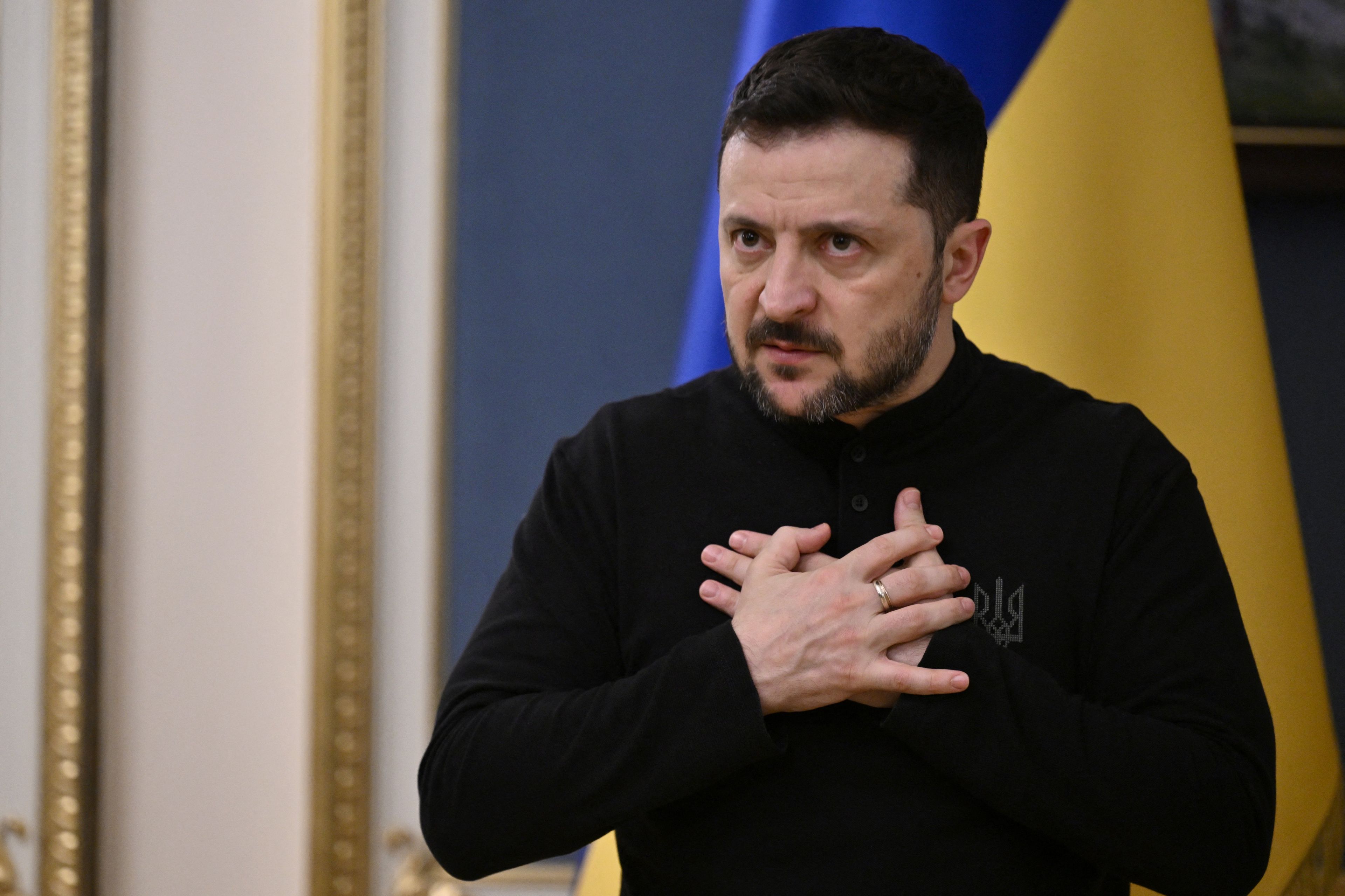 Na zdjęciu jest mężczyzna ok. 45 lat. Ma czarne włosy i krótką brodę. Jest w czarnej bluzie. Z tyłu jest flaga o kolorach niebiesko-żółtych