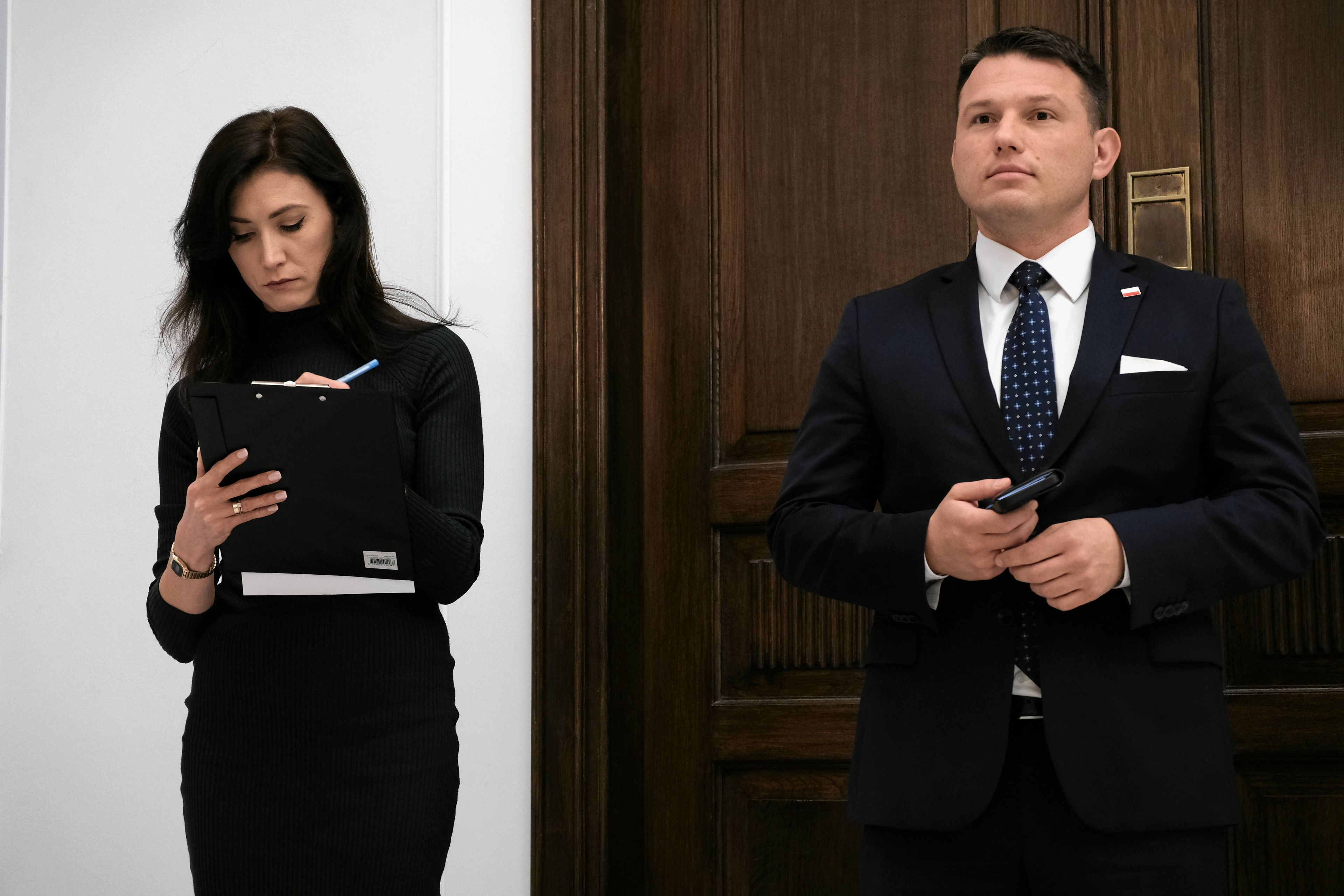 Mężczyzna i kobieta stoją na tle białej ściany i ciemnych drzwi. Kobieta ma czarne długie włosy i czarną obcisłą sukienkę. Mężczyzna jest ubrany w granatowy garnitur i białą koszulę.