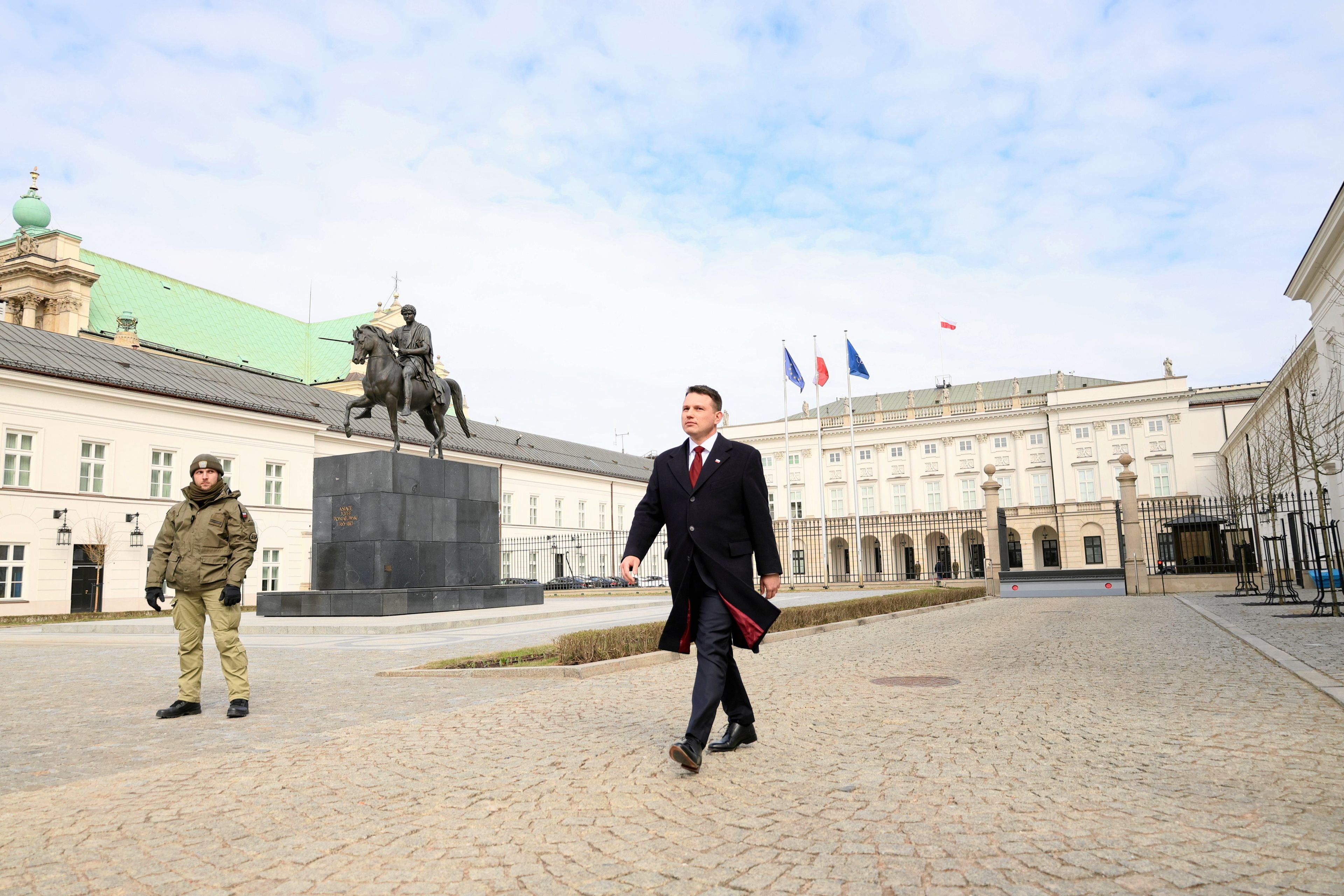 Mężczyzna w ciemnym płaszczu idzie po dziedzińcu Pałacu Prezydenckiego w stronę bramy.