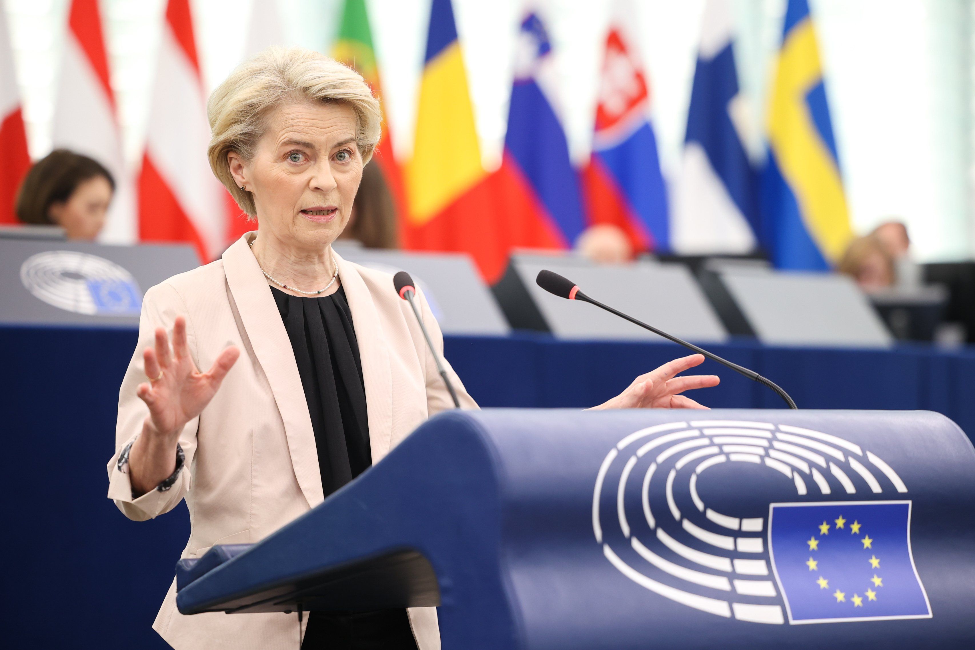 Kobieta, blondynka z krótkimi włosami bez grzywki założonymi za uszy, stoi przy niebieskiej mównicy i gestykuluje rękami. Jej wyraz twarzy sugeruje stanowczość i podkreślenie czegoś ważnego. W tle kilka kolorowych flag państw unijnych i niebieskie pulpity, przy których siedzą osoby przewodniczące spotkaniu. Kobieta na pierwszym planie ma na sobie kremową, krótką marynarkę i czarną bluzkę.