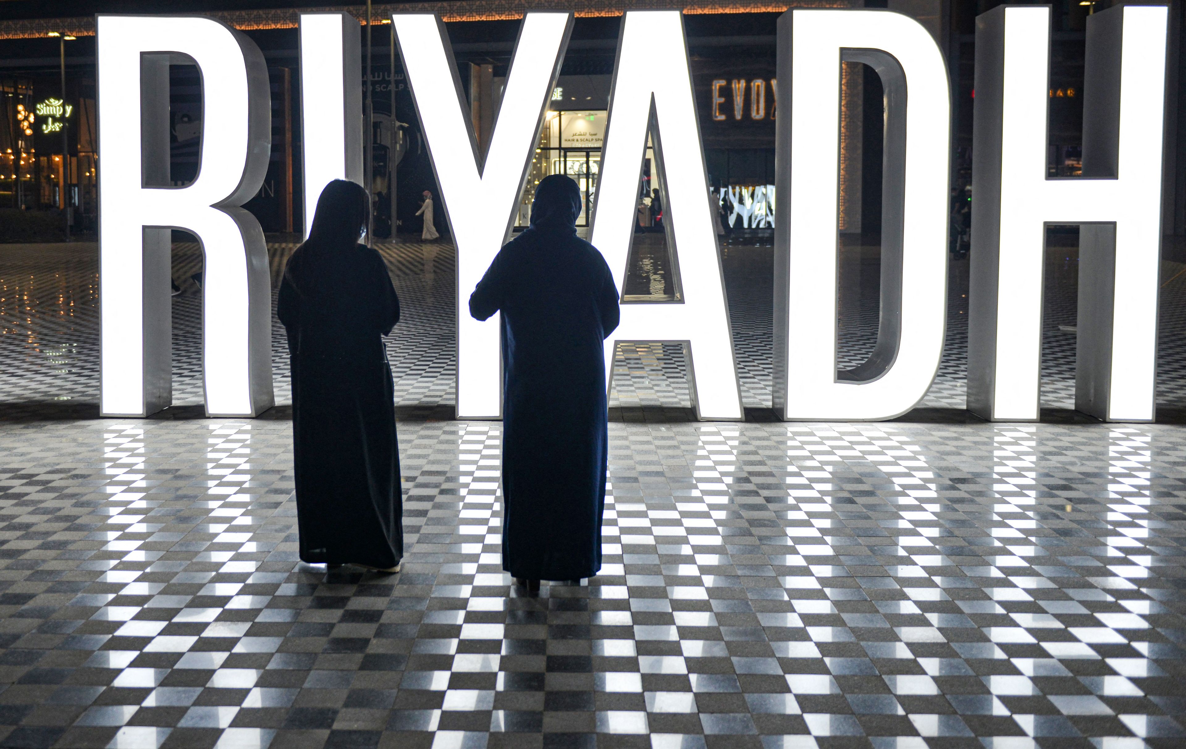 Dwie kobiety w abajach i hidżabach stoją przed wielkim napisem RIYADH.