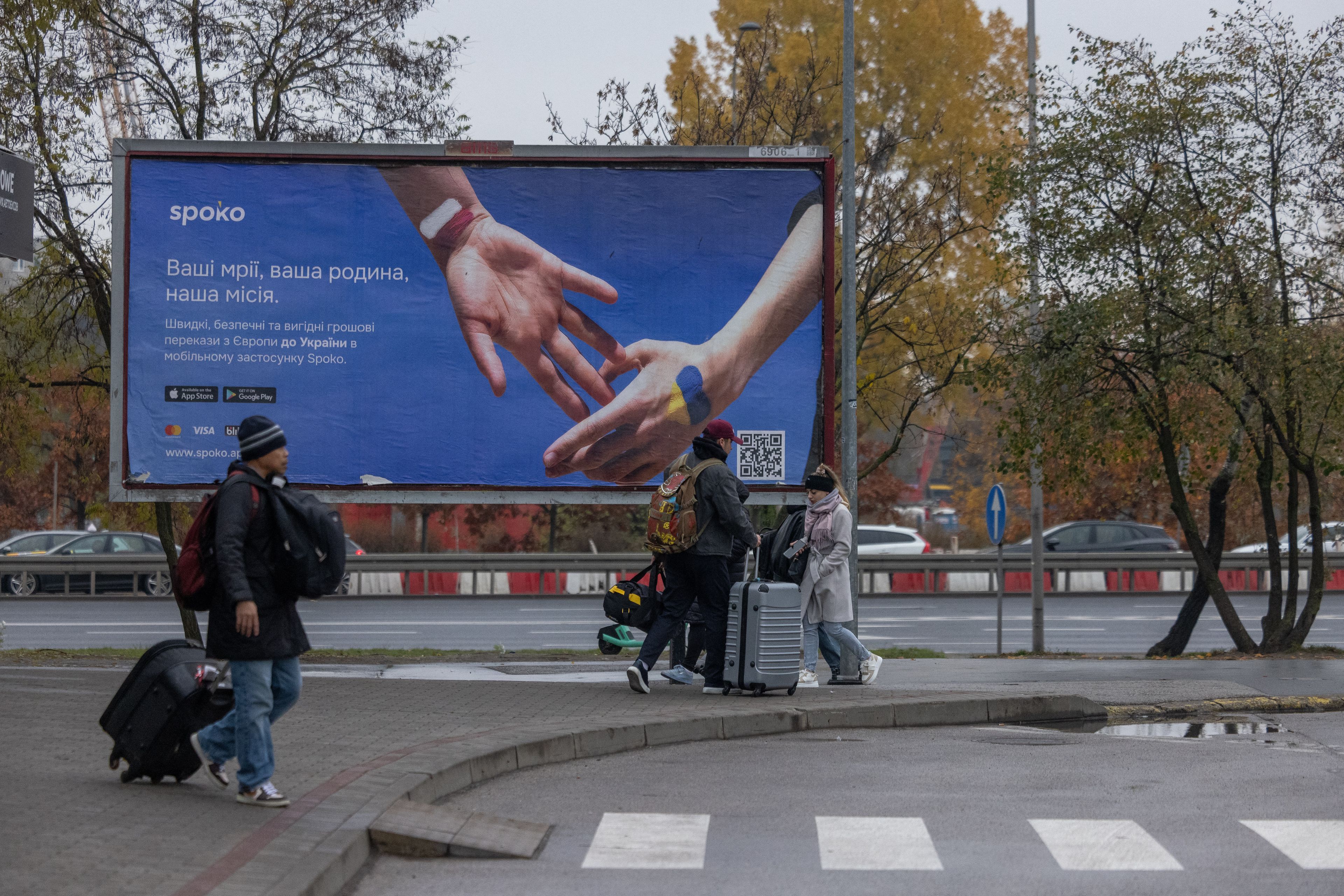 Travellers pass by a poster of a company transferring money from Poland to Ukraine at Warsaw West bus station on October 30, 2024. War, interrogations and a journey lasting for days are not enough to stop thousands of Ukrainians from a massive round-about circuit from Poland to the occupied Ukraine through Russia, going through intense checks set up in Moscow (Photo by Wojtek RADWANSKI / AFP)