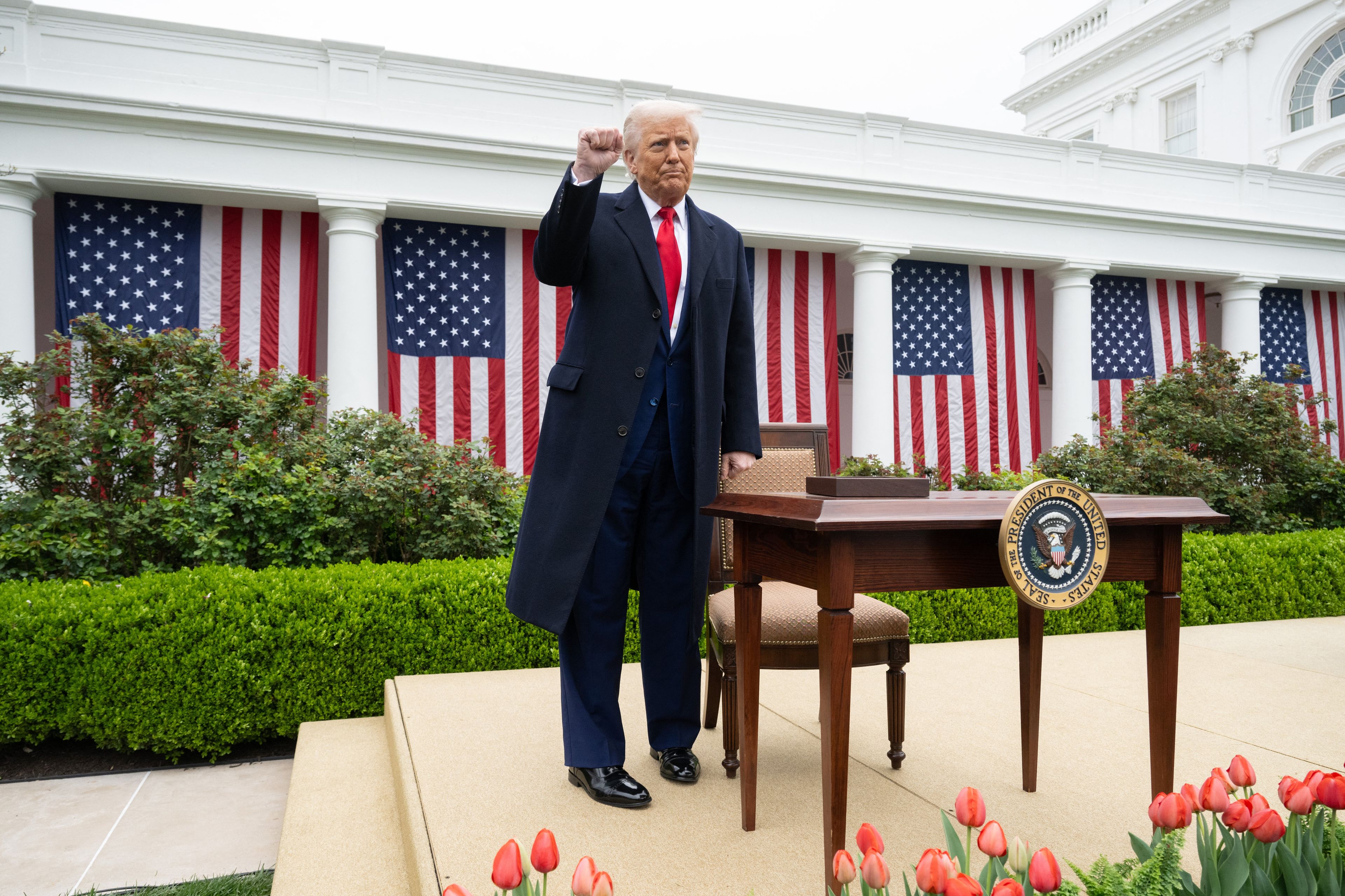 Donald Trump przed Białym Domem obok stolika, stoi z wyciągniętą ręką w geście triumfu
