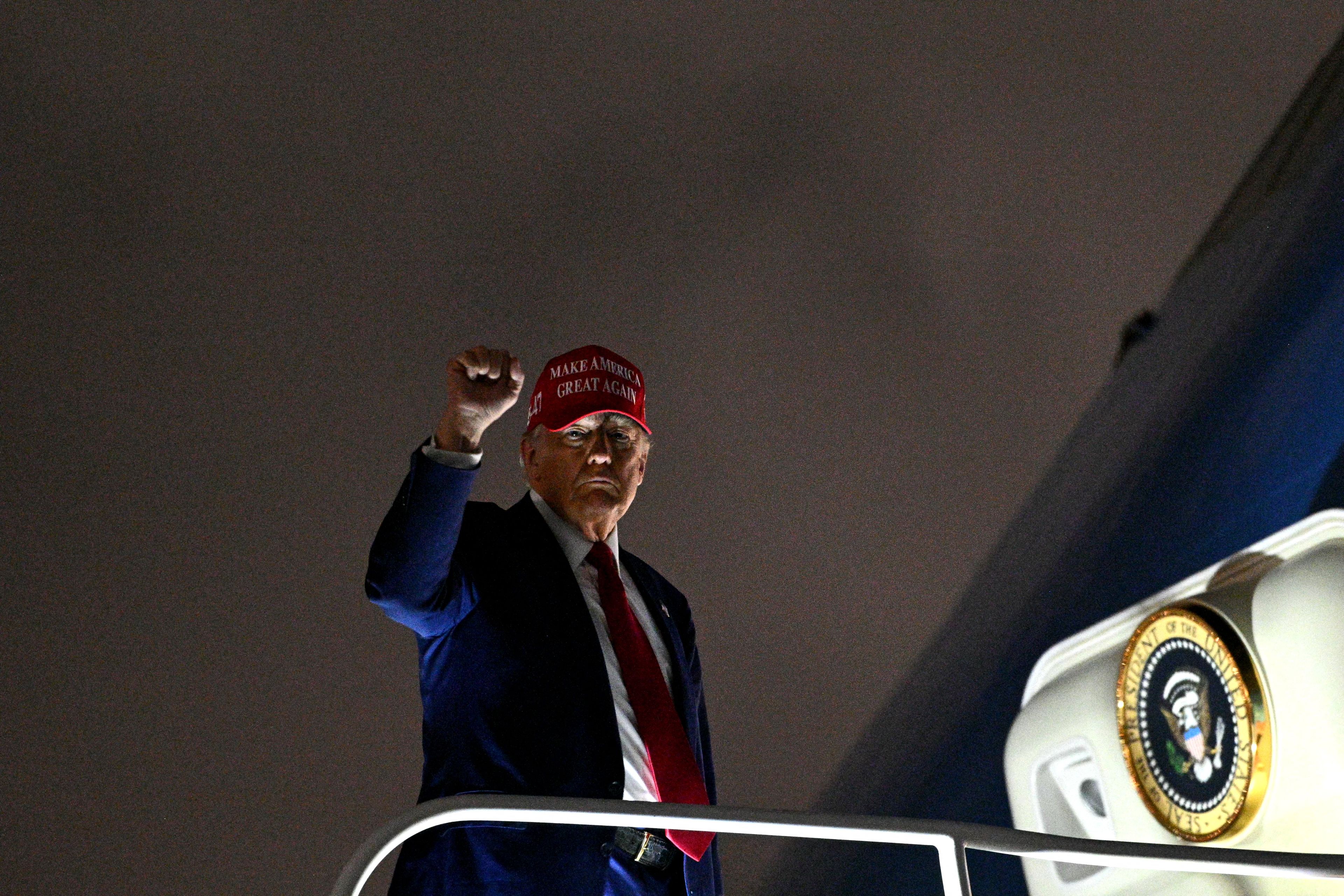 Donald Trump w czerwonej czapce wchodzi na pokład samolotu, unosi dłoń w górę w geście tryiumfu
