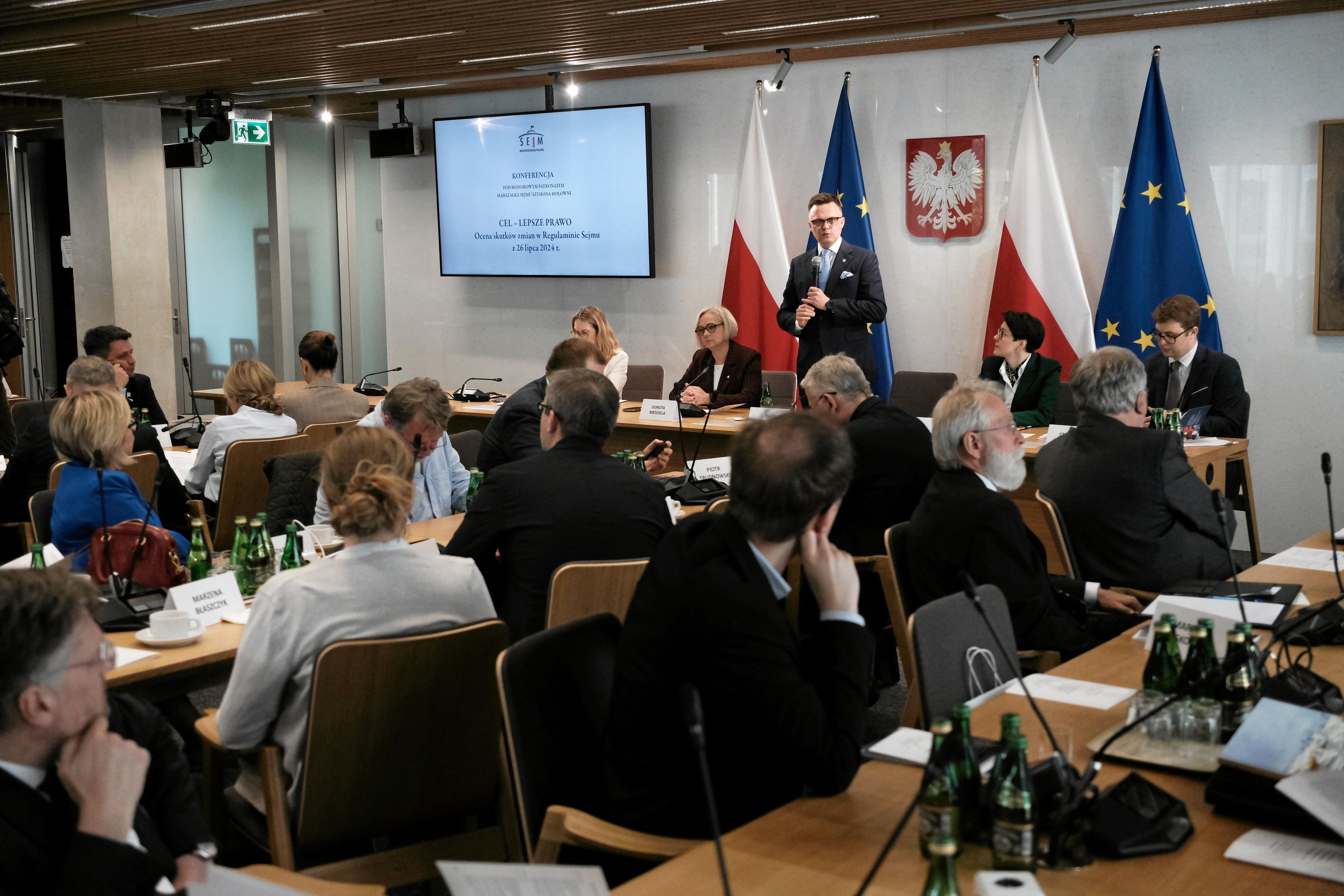 Męzczyzna w garniturze przemawia stojąc w sali pełnej ludzi