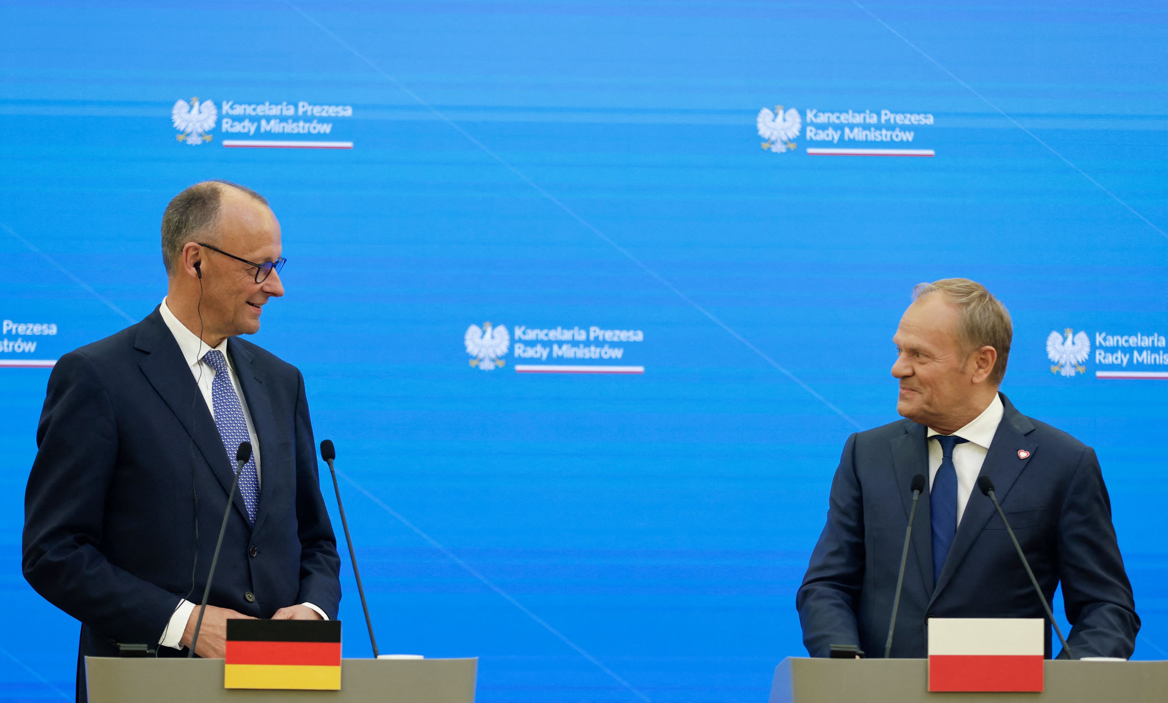 Dwóch mężczyzn w garniturach stoi obok siebie przy mównicach podczas konferencji prasowej. Mężczyzna po lewej stronie ma okulary, jest łysy, ubrany w ciemny garnitur, białą koszulę i niebieski krawat w drobny wzór; nosi słuchawkę w uchu. Mężczyzna po prawej ma jasne włosy zaczesane do tyłu, ubrany jest w granatowy garnitur, białą koszulę i granatowy krawat; na klapie ma przypinkę w kształcie serca. Obaj uśmiechają się do siebie. W tle niebieska ściana z emblematem "Kancelaria Prezesa Rady Ministrów", a przed nimi mównice z flagami Niemiec i Polski.