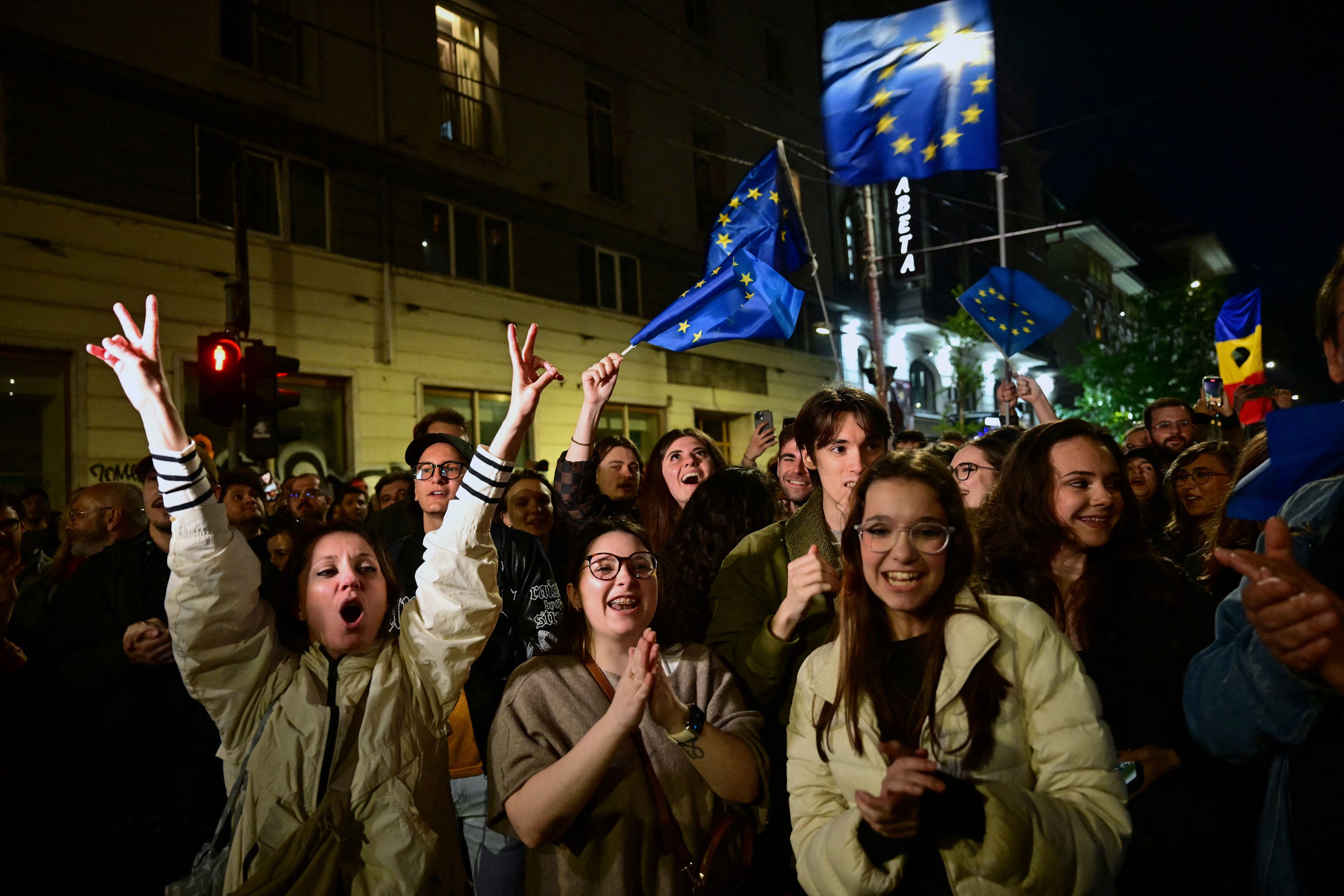 Grupa młodych ludzi zgromadzona nocą na ulicy świętuje zwycięstwo wyborcze swojego faworyta Nicușora Dana. Uczestnicy uśmiechają się, klaszczą i unoszą ręce w geście zwycięstwa. Wśród nich dominują kobiety i mężczyźni w wieku około 20–30 lat, ubrani swobodnie, niektórzy z okularami. W tle powiewają liczne flagi Unii Europejskiej, a także flaga Rumunii. Otoczenie to miejska ulica – widać budynki z oknami, neon z napisem „Libertă” i oświetlenie uliczne. Całość emanuje entuzjazmem, zaangażowaniem obywatelskim i atmosferą celebracji demokratycznego sukcesu.