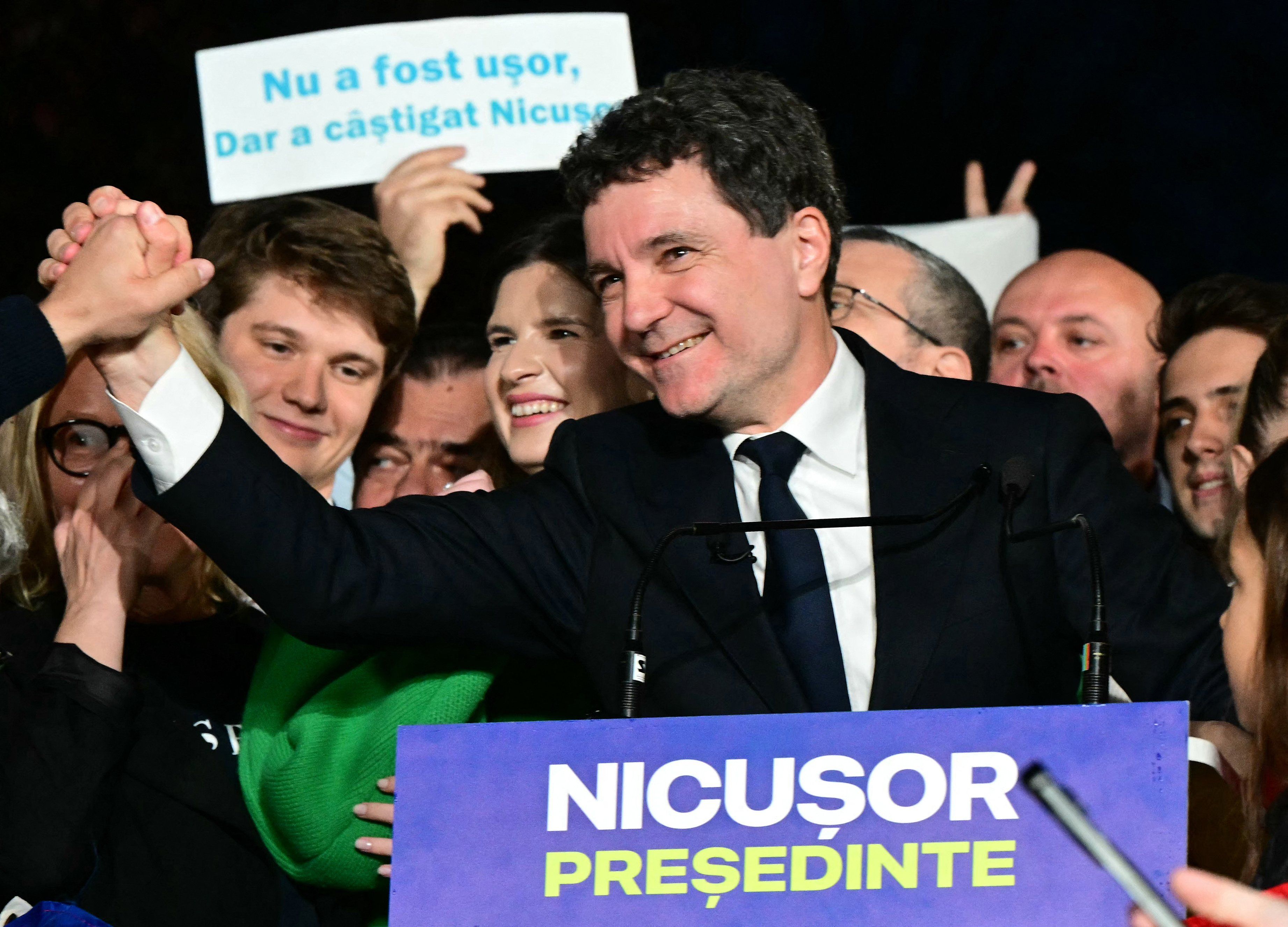Mężczyzna w średnim wieku, ubrany w ciemny garnitur, biały podkoszulek i granatowy krawat, uśmiecha się szeroko, ściskając rękę innej osoby na scenie w geście zwycięstwa. W tle widać wiwatujący tłum jego zwolenników – młodych i uśmiechniętych ludzi, z których część trzyma transparenty. Jeden z nich głosi: „Nu a fost ușor, dar a câștigat Nicușor” („Nie było łatwo, ale Nicușor wygrał”). Zdjęcie ma charakter dynamiczny, emocjonalny i celebracyjny.
