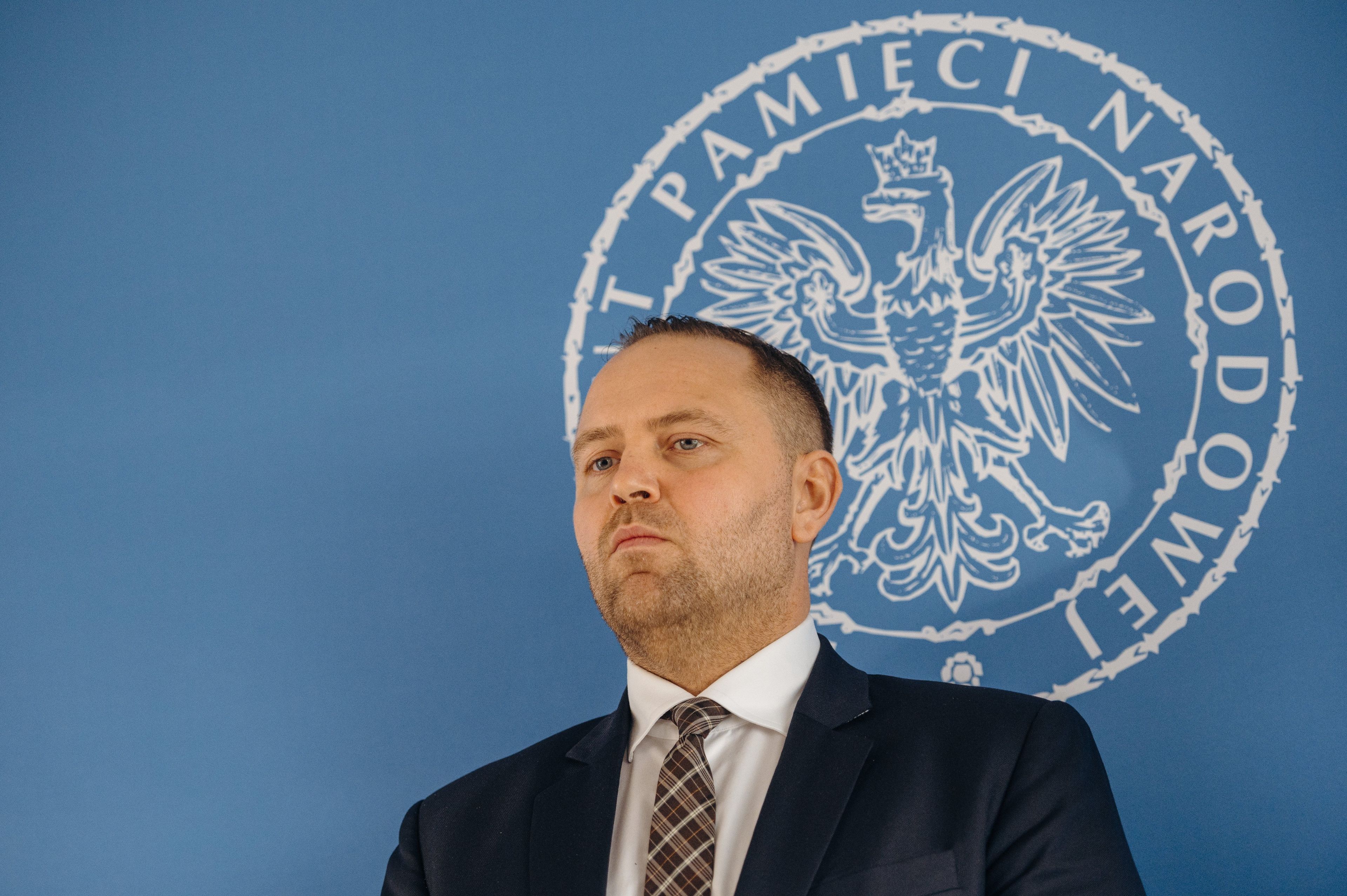Mężczyzna w garniturze na tle niebieskiej ścianki z logiem IPN