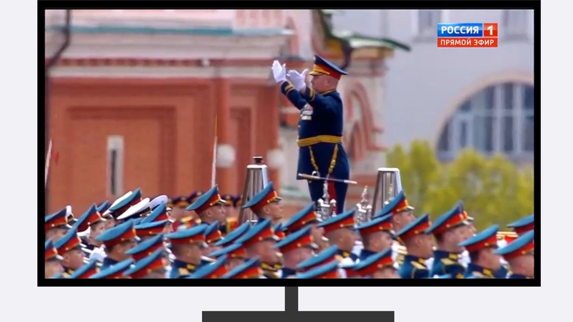 Grubszy wojskowy w galowym mundurze dyryguje z podwyższenia orkiestrą wojskową. Poniżej widać głowy żołnierzy. Kadr z telewizji wstawiony w ramkę telewizyjnego ekranu