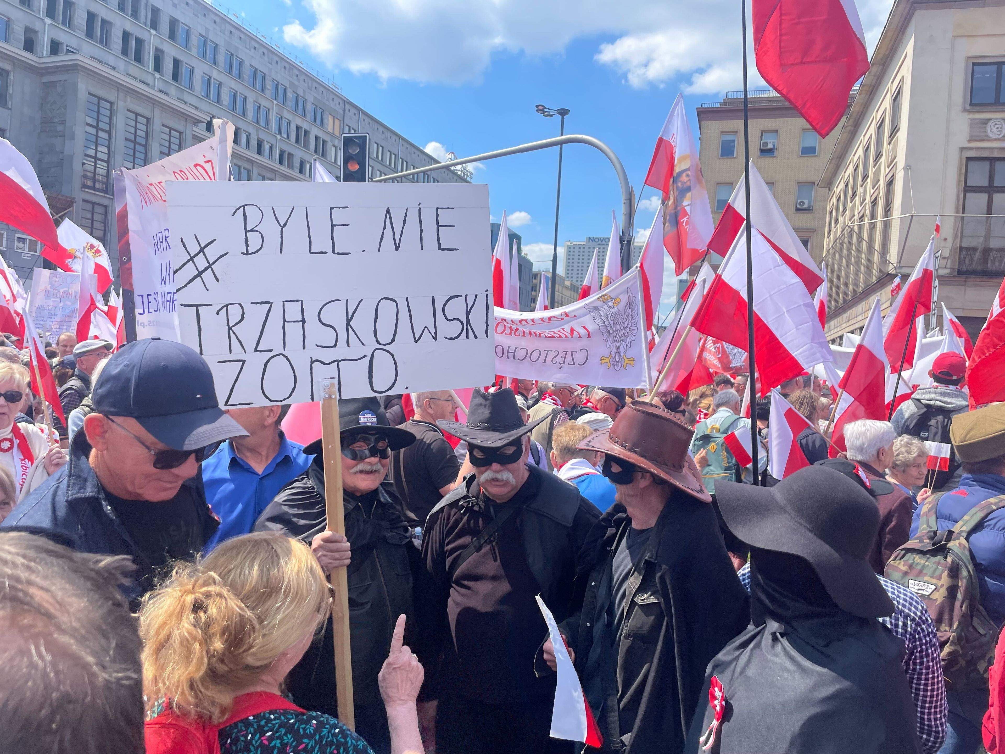 W tłumie stoi trzech mężczyzn ubranych na czarno, z czarnymi przepaskami na oczach i czarnymi kapeluszami na głowach. Trzymają transparent z napisem "Byle nie Trzaskowski, Zorro"