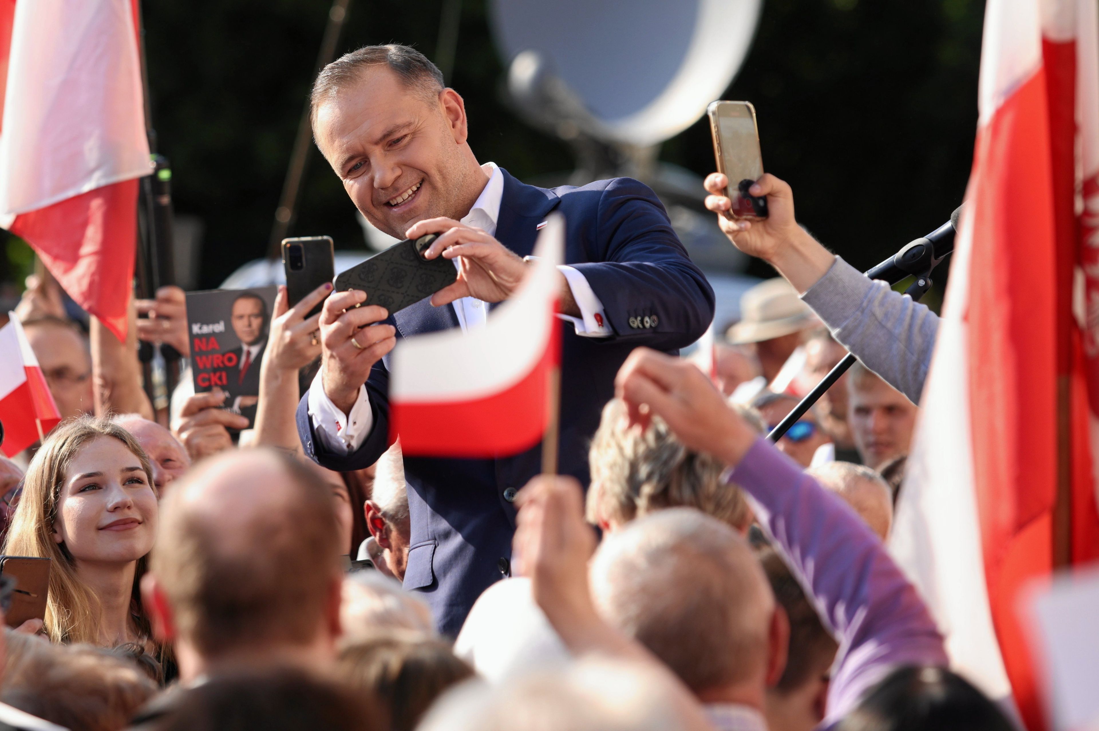 Uśmiechnięty 40-latek w garniturze przed tłumem wiwatujących ludzi z biało-czerwonymi flagami.