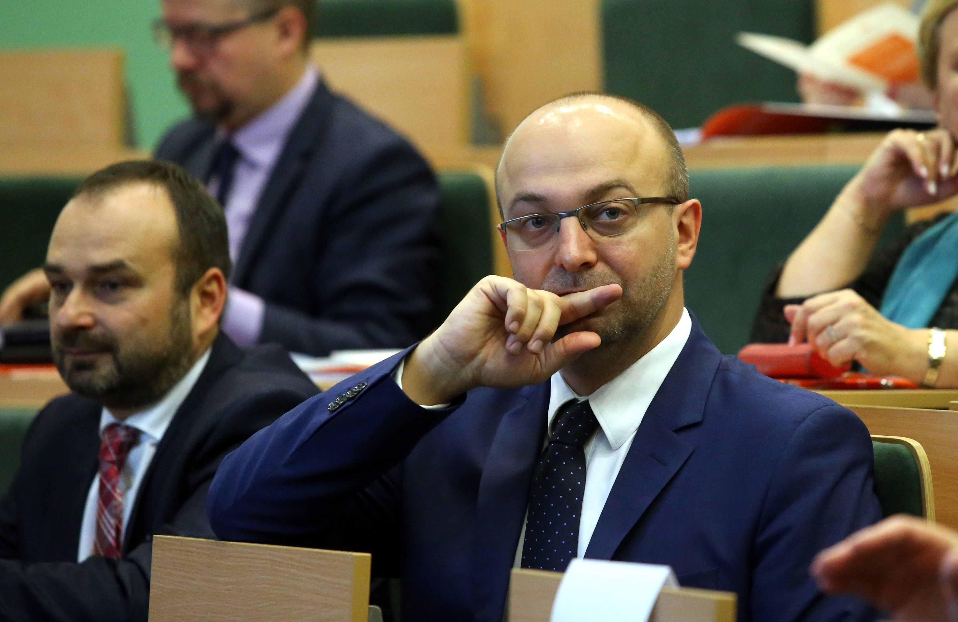 Łysy mężczyzna w sednim wieku, w okularach na sali, gdzie jest wokół niego dużo osób