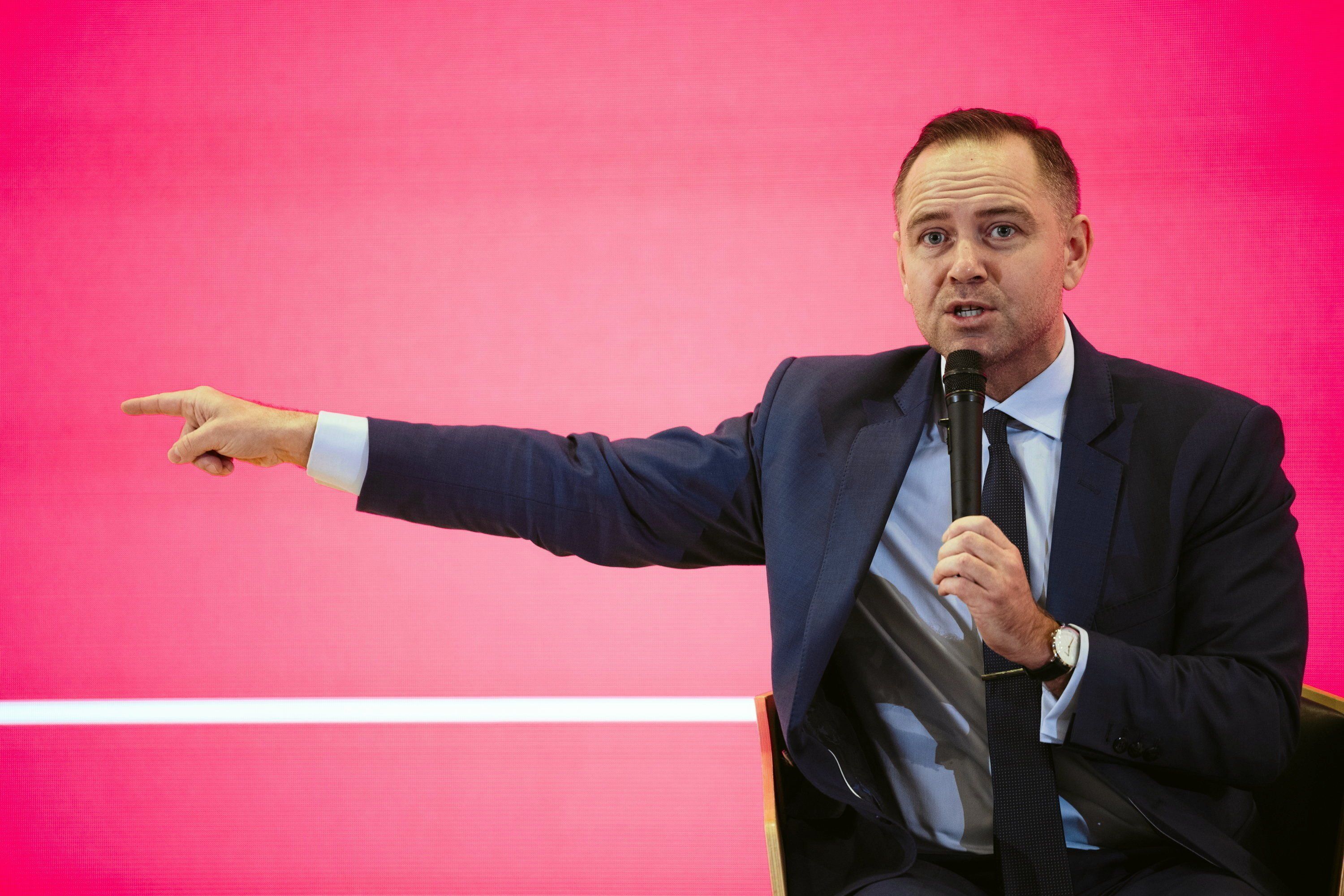 Mężczyzna w garniturze z mikrofonem w ręku przemawia wskazując ręką w prawo