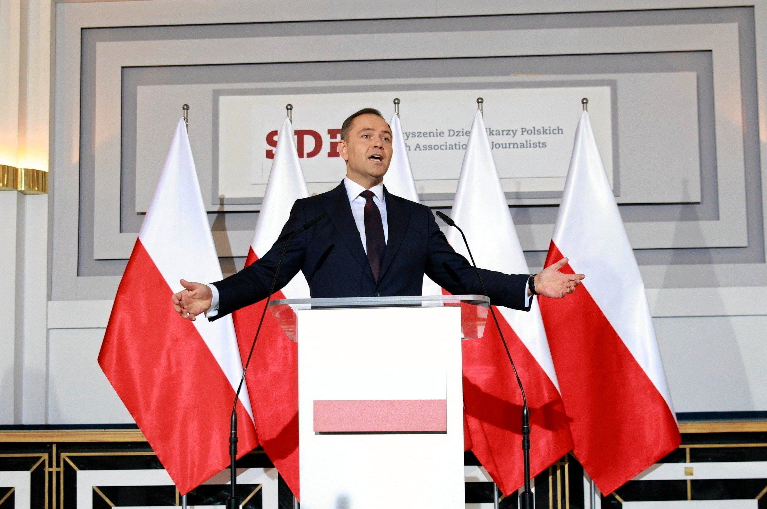 W dużej sali stoi przed mikrofonem mężczyzna i rozkłada szeroko ręce w geście